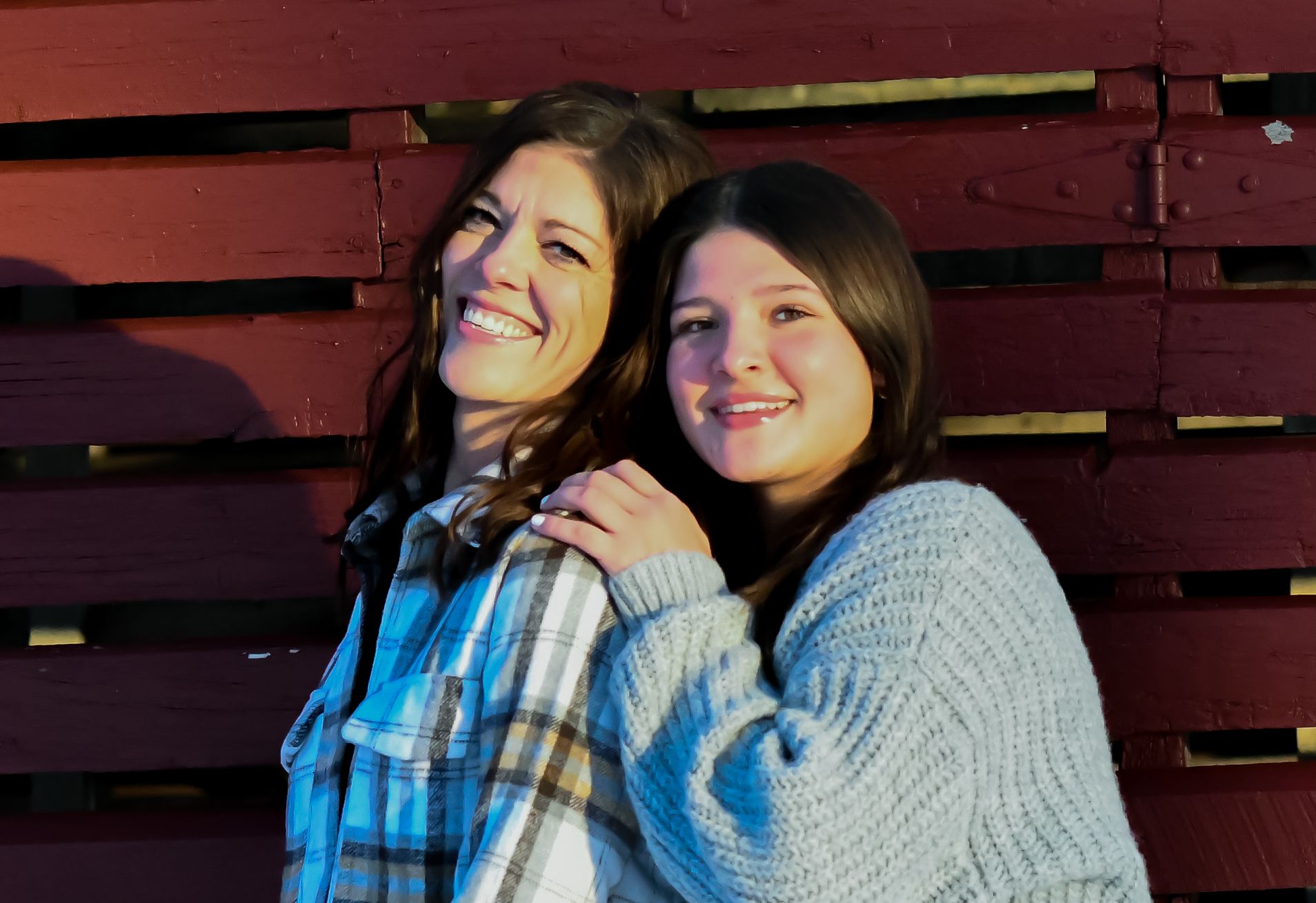 Two smiling women pose together in front of a red wooden fence, wearing casual flannel and knit sweaters.