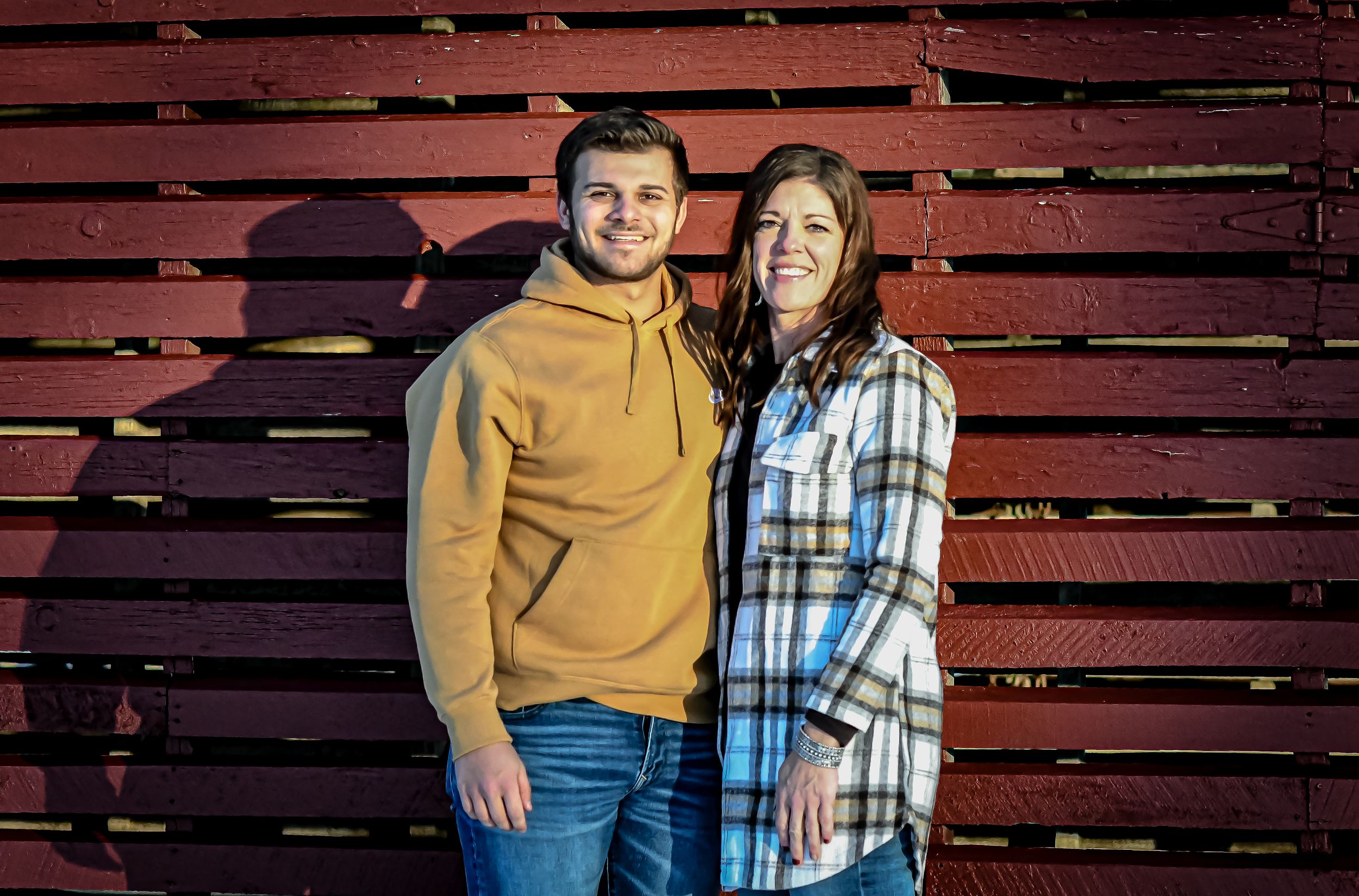 A young man in a yellow hoodie and a woman in plaid shirt smile together against a rustic red wooden wall.