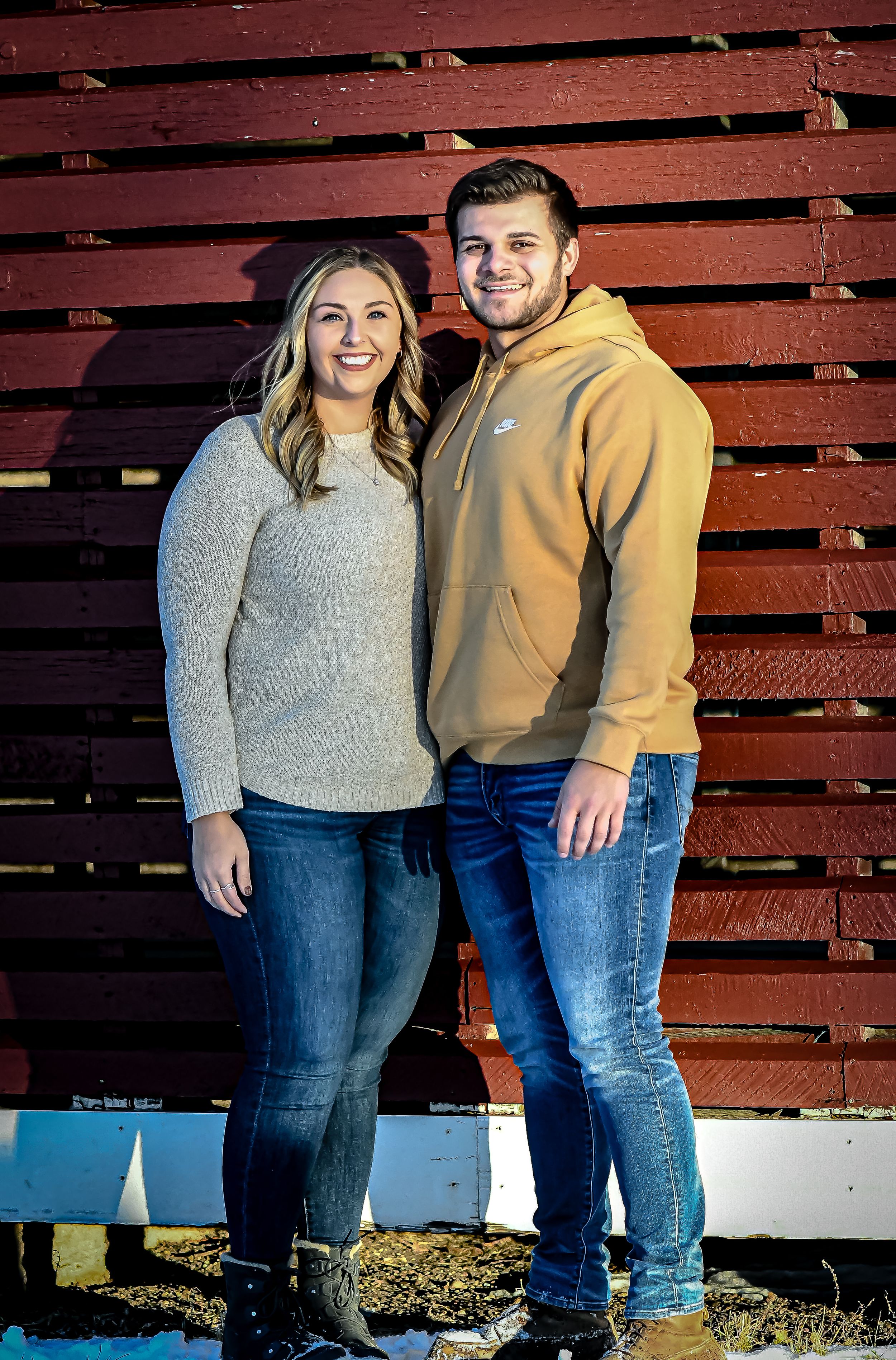 Smiling couple posing together in front of a red barn wall, wearing casual sweater and hoodie.