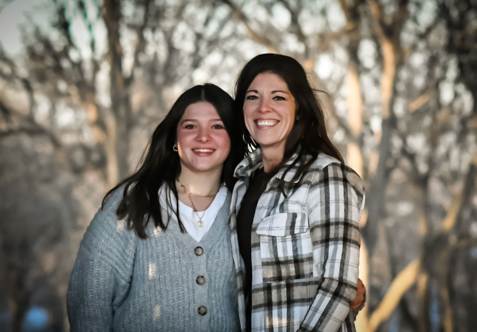 Two smiling women pose together outdoors in winter, wearing cozy sweaters with bare trees in the background.