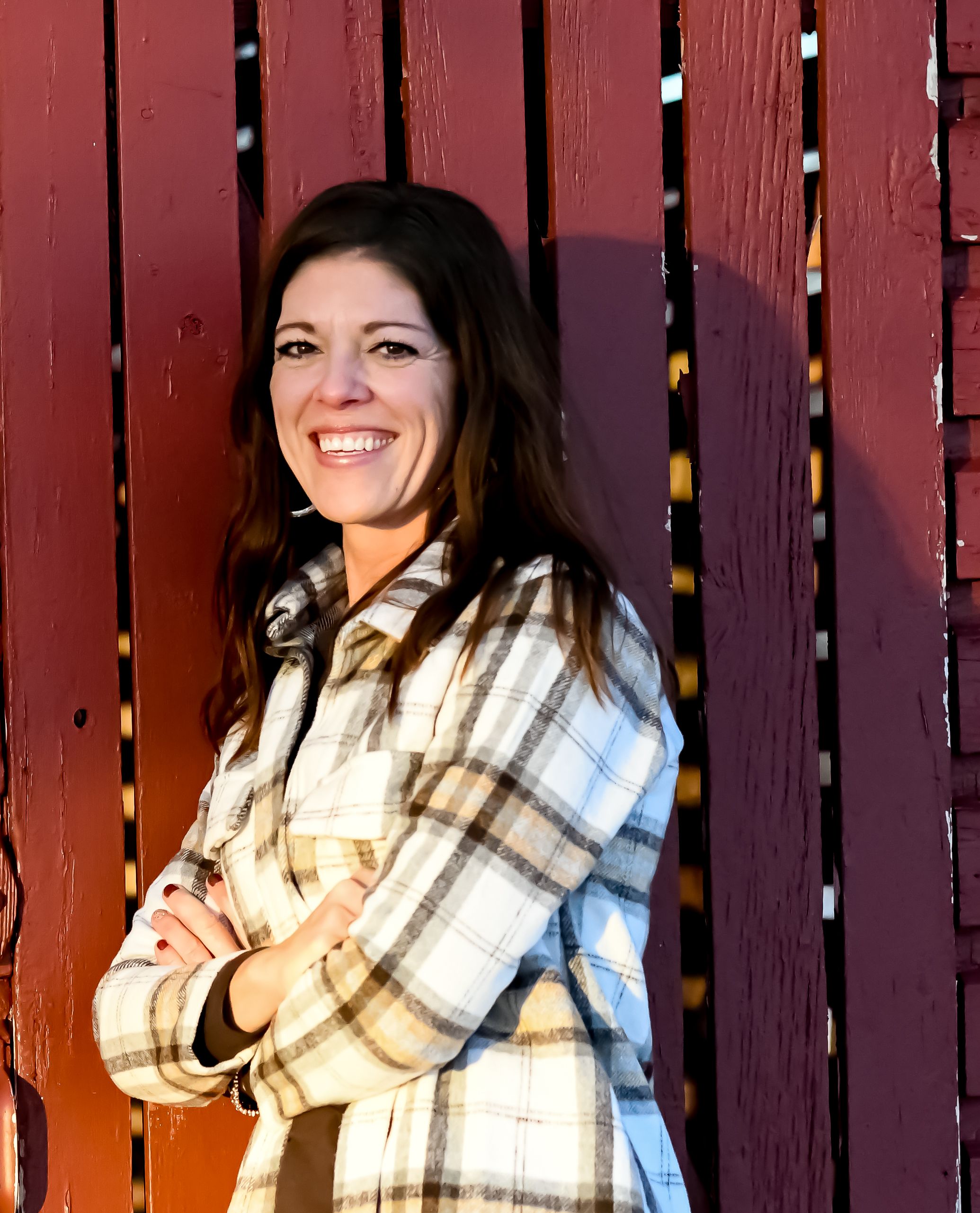 Smiling woman with crossed arms in plaid shirt leaning against red wooden fence in sunny outdoor setting.