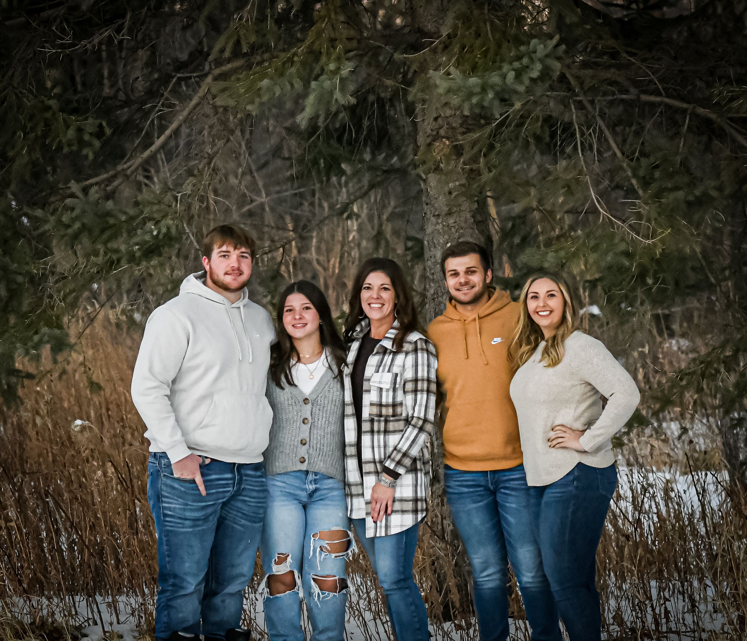 Five people pose together outdoors in winter, smiling in front of snow-covered ground and pine trees.