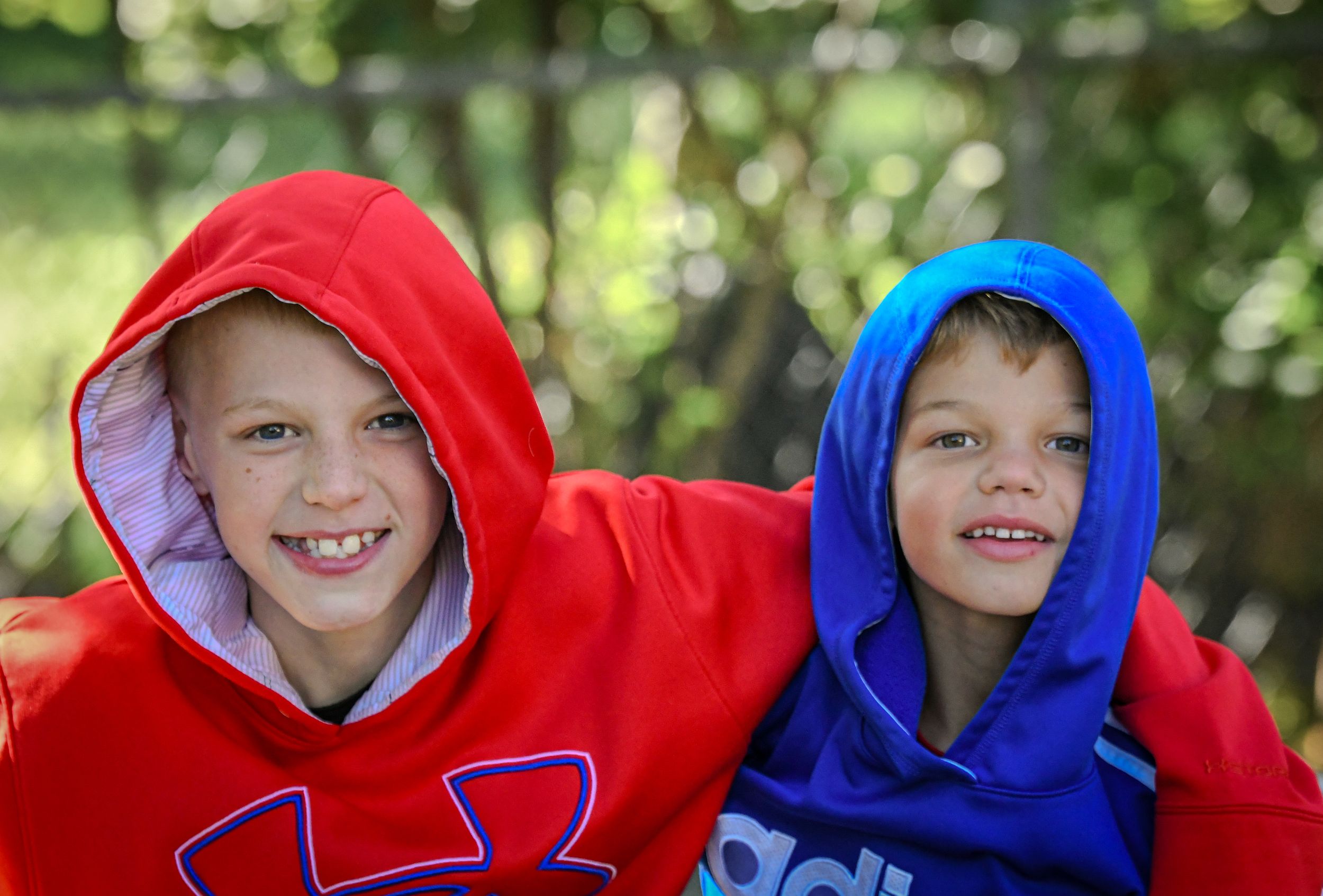 Two smiling children wearing colorful hoodies, one red and one blue, posing together outdoors with green trees in background.