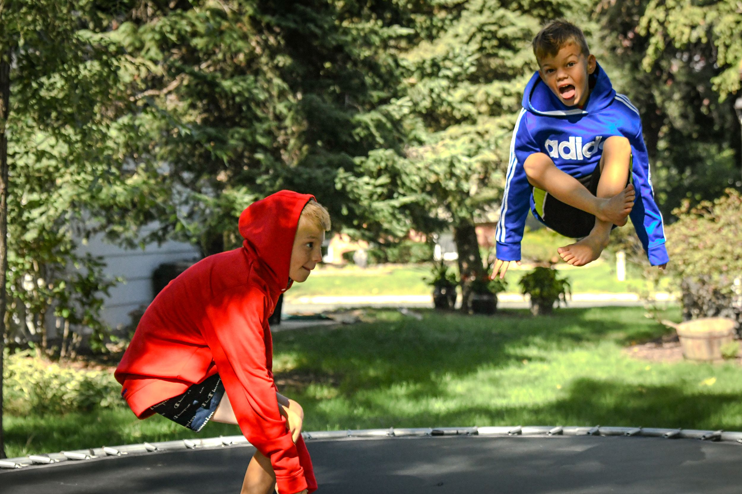 Two kids jumping on a trampoline outdoors, one in red and one in a blue Adidas hoodie, surrounded by green trees.