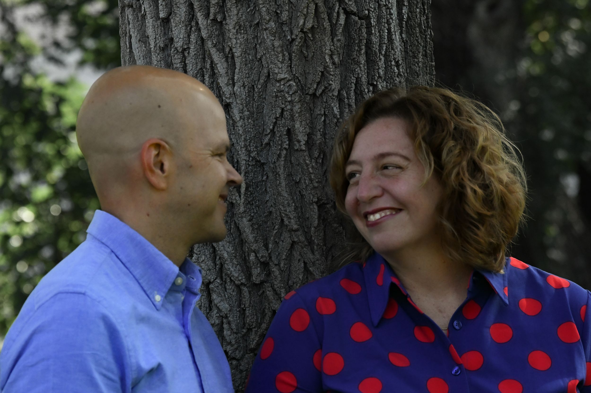 A smiling couple leans against a tree outdoors, the woman in a red polka dot blouse and the man in a blue shirt.