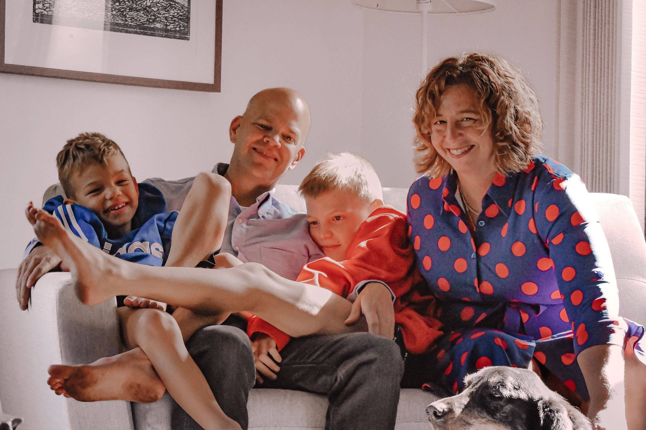 Happy family of four with two young boys sitting together on a white sofa at home, smiling warmly.
