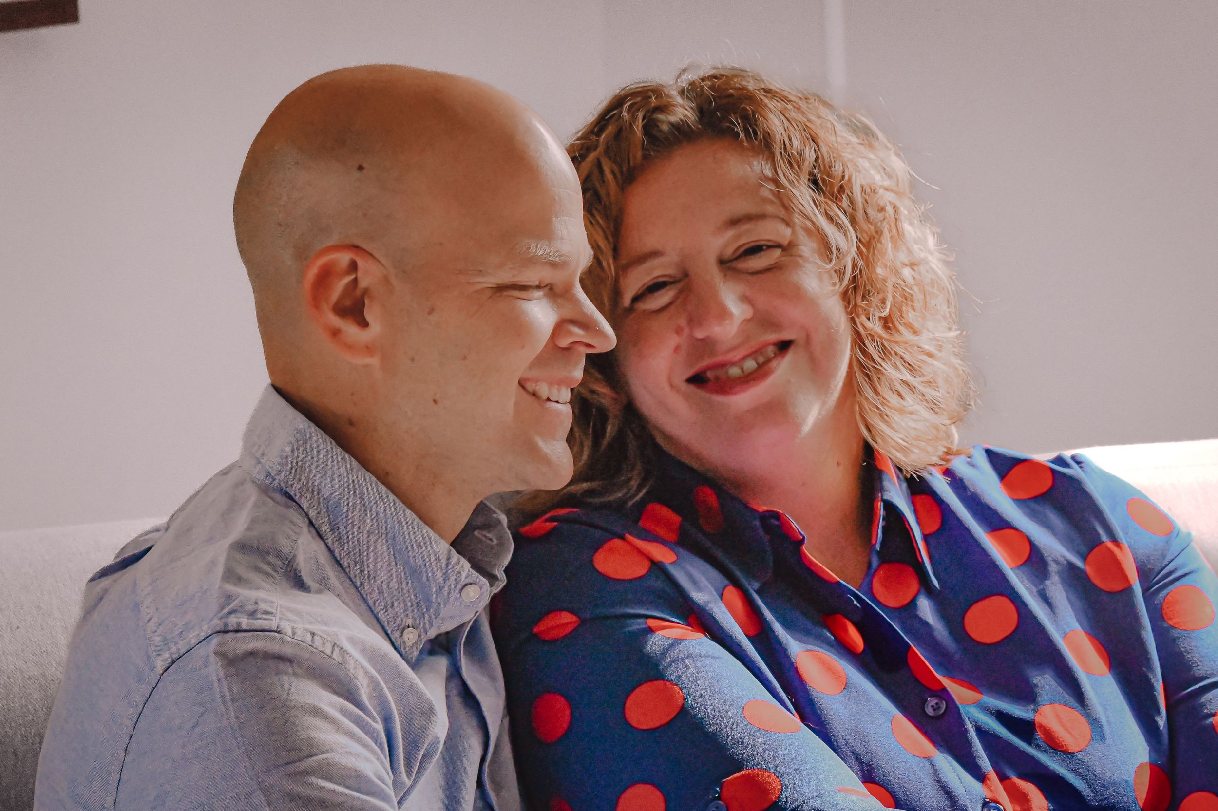 A smiling bald man and curly-haired woman in a blue polka dot shirt share a warm moment together on a sofa.