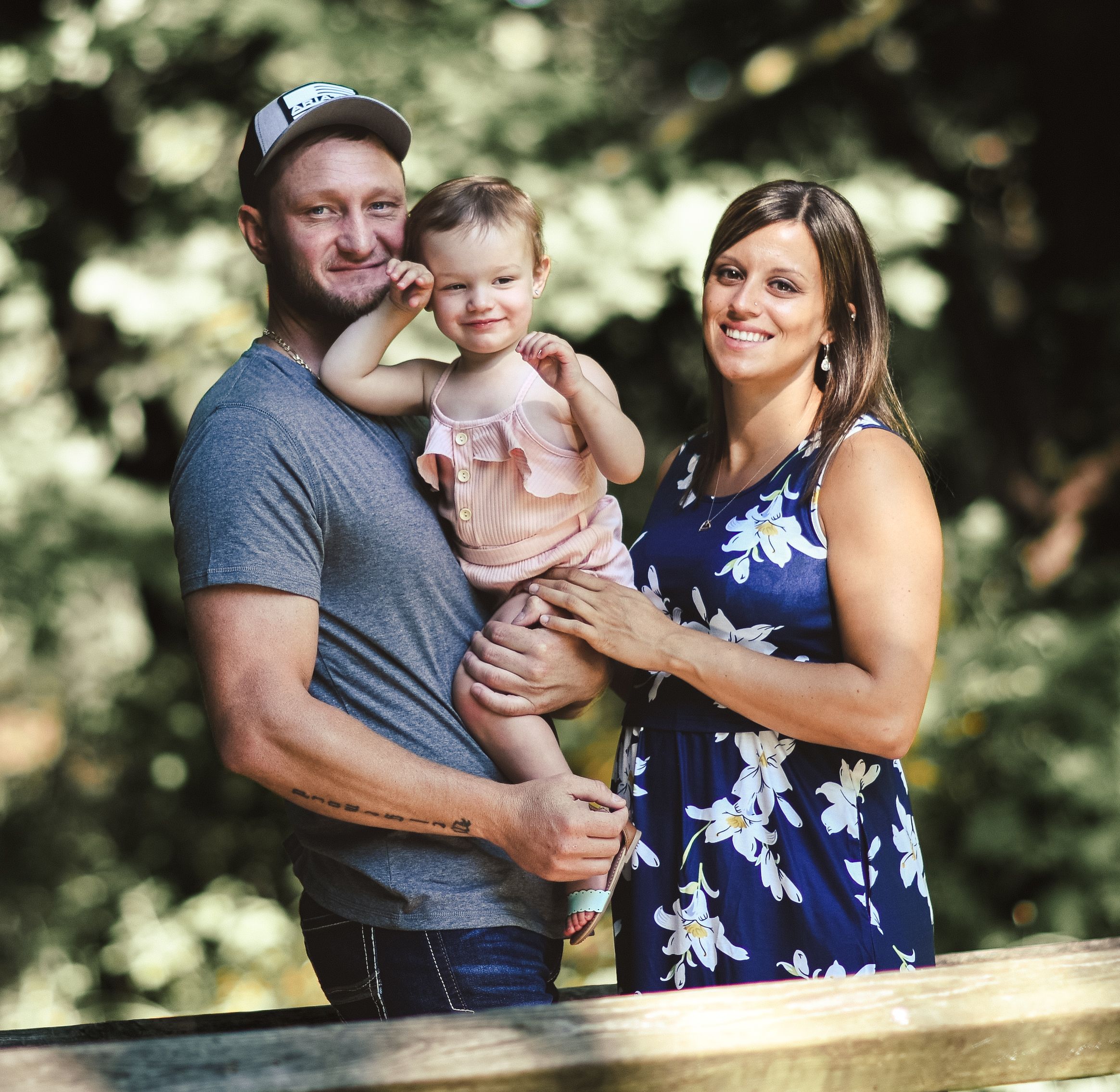 Happy young family with toddler posing outdoors among lush green trees in a warm summer portrait.
