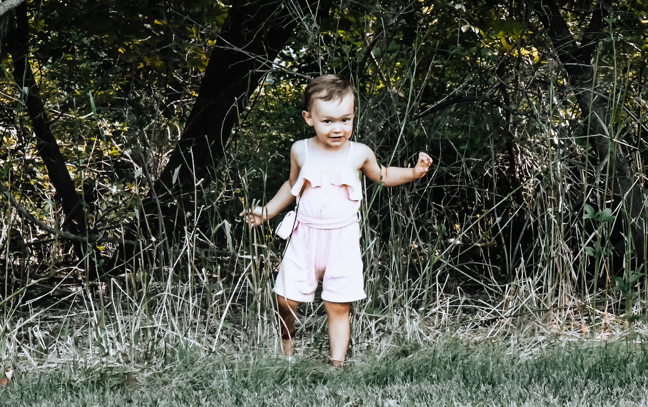 A toddler in a pink outfit walks through tall grass near trees in a sunny outdoor setting.