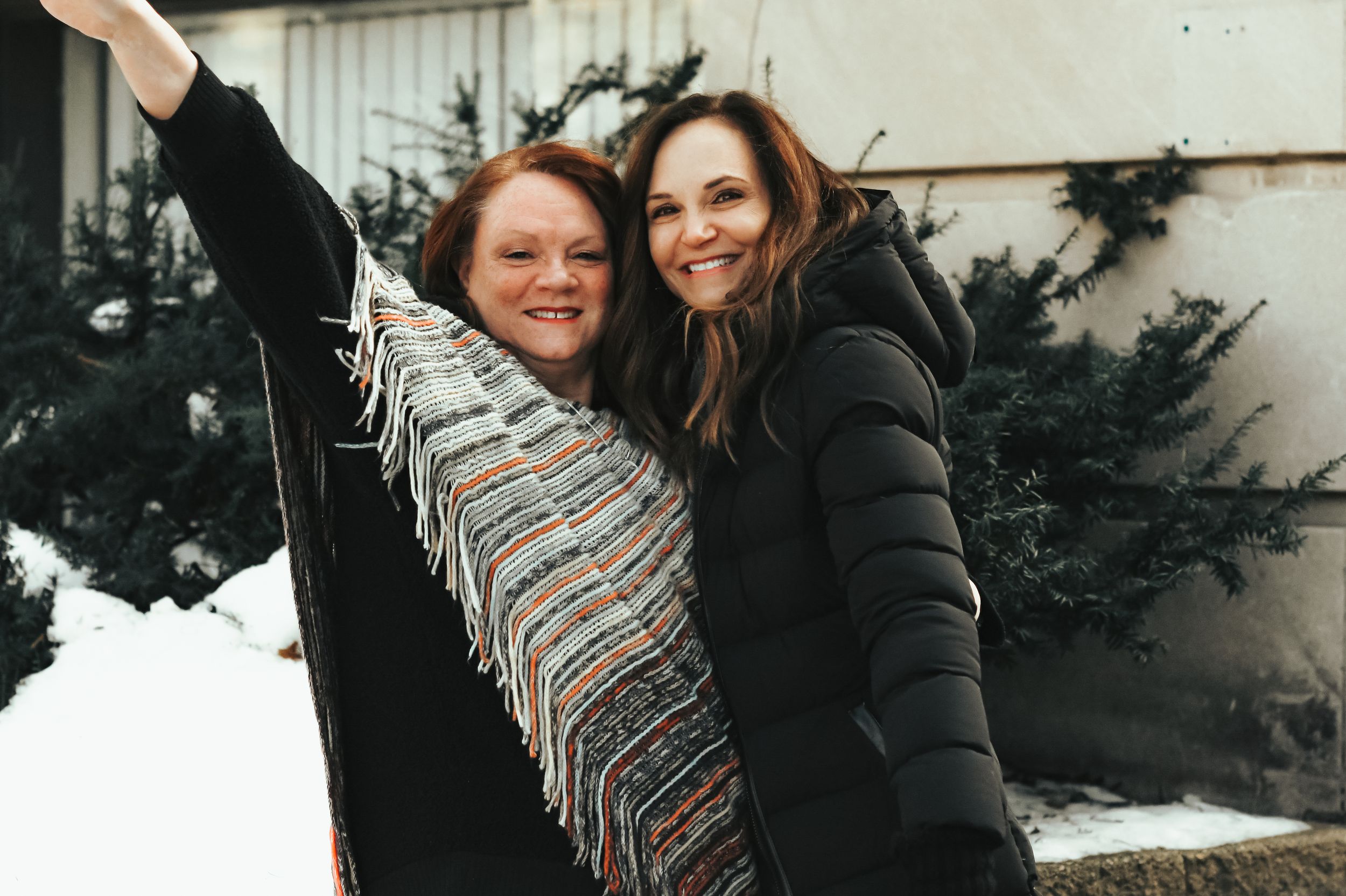 Two women smiling and embracing outdoors in winter, one wearing a colorful scarf, with snow and evergreen trees behind them.