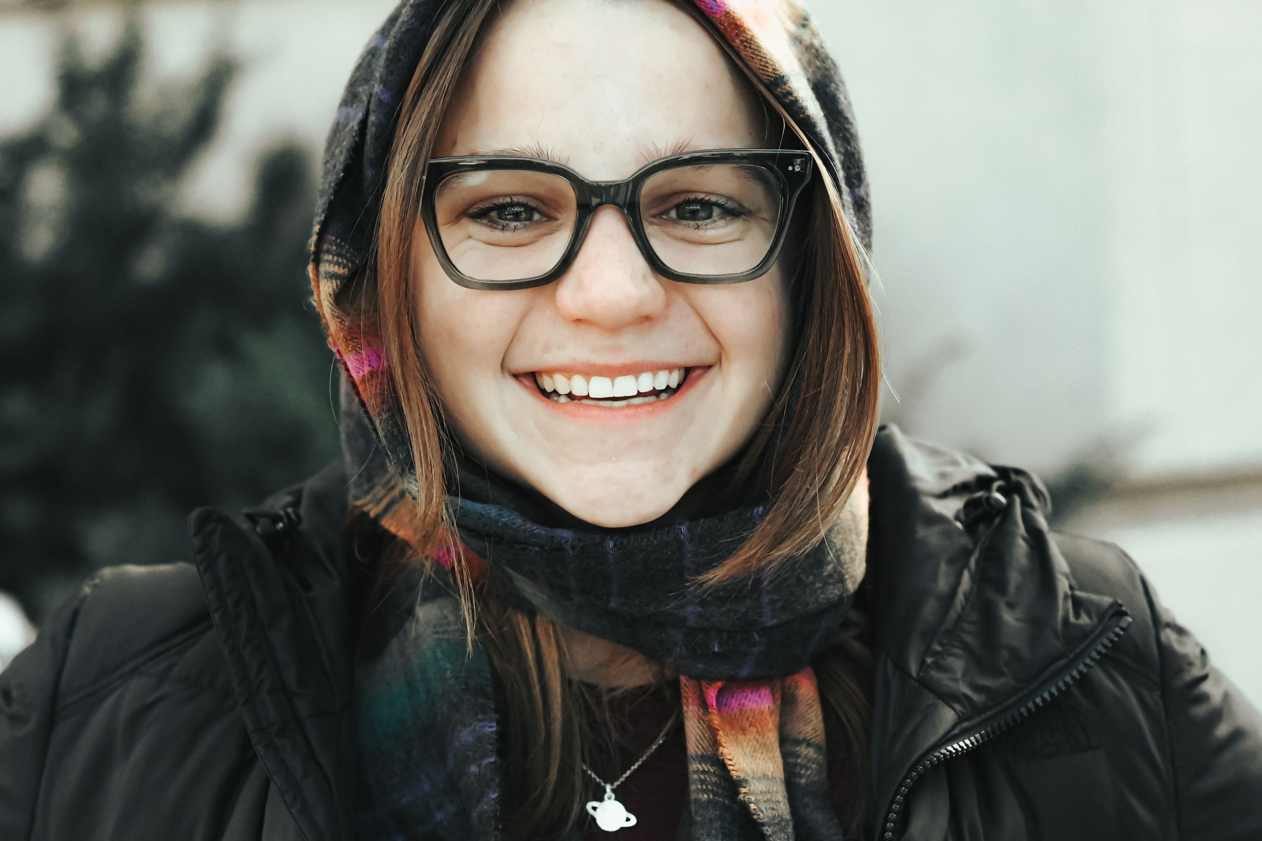 Smiling young woman wearing black glasses, colorful scarf, and hooded jacket outdoors in winter.