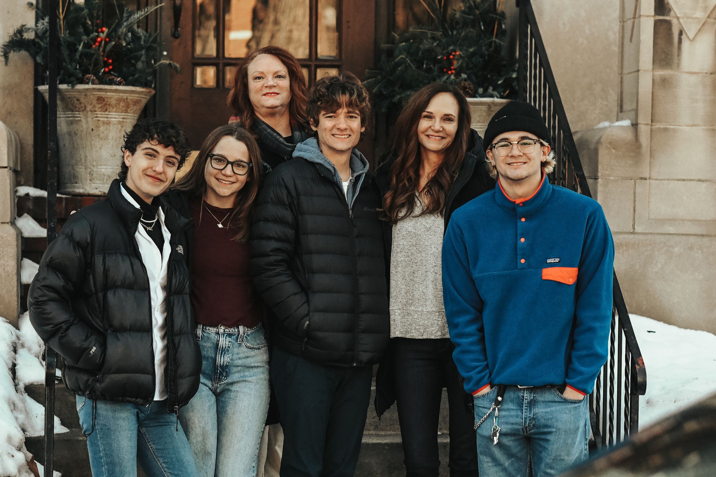 Six people smiling together outdoors on snowy steps, wearing winter clothing for a casual family group photo.
