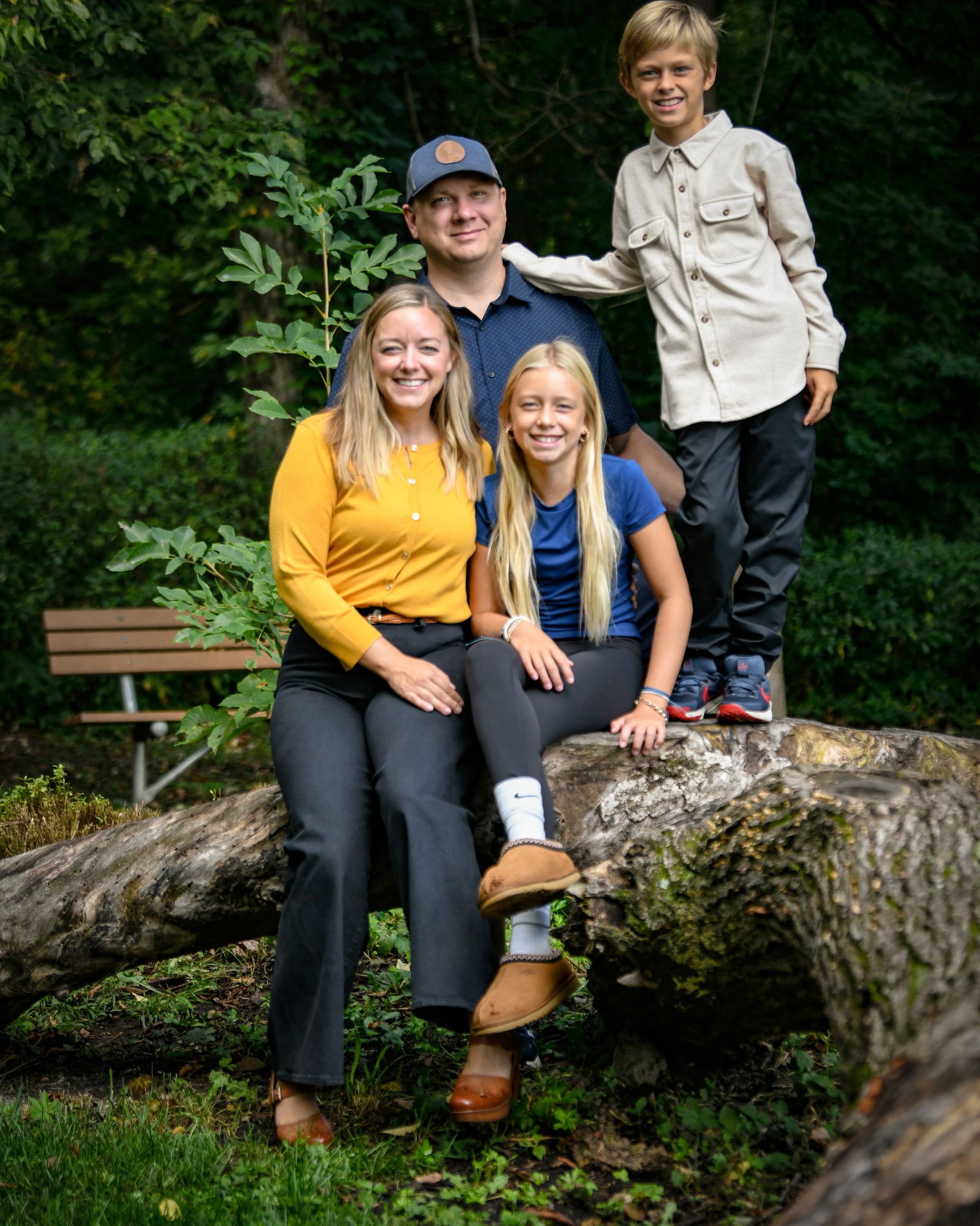 A smiling family of four poses outdoors on a fallen log surrounded by lush green trees and foliage.
