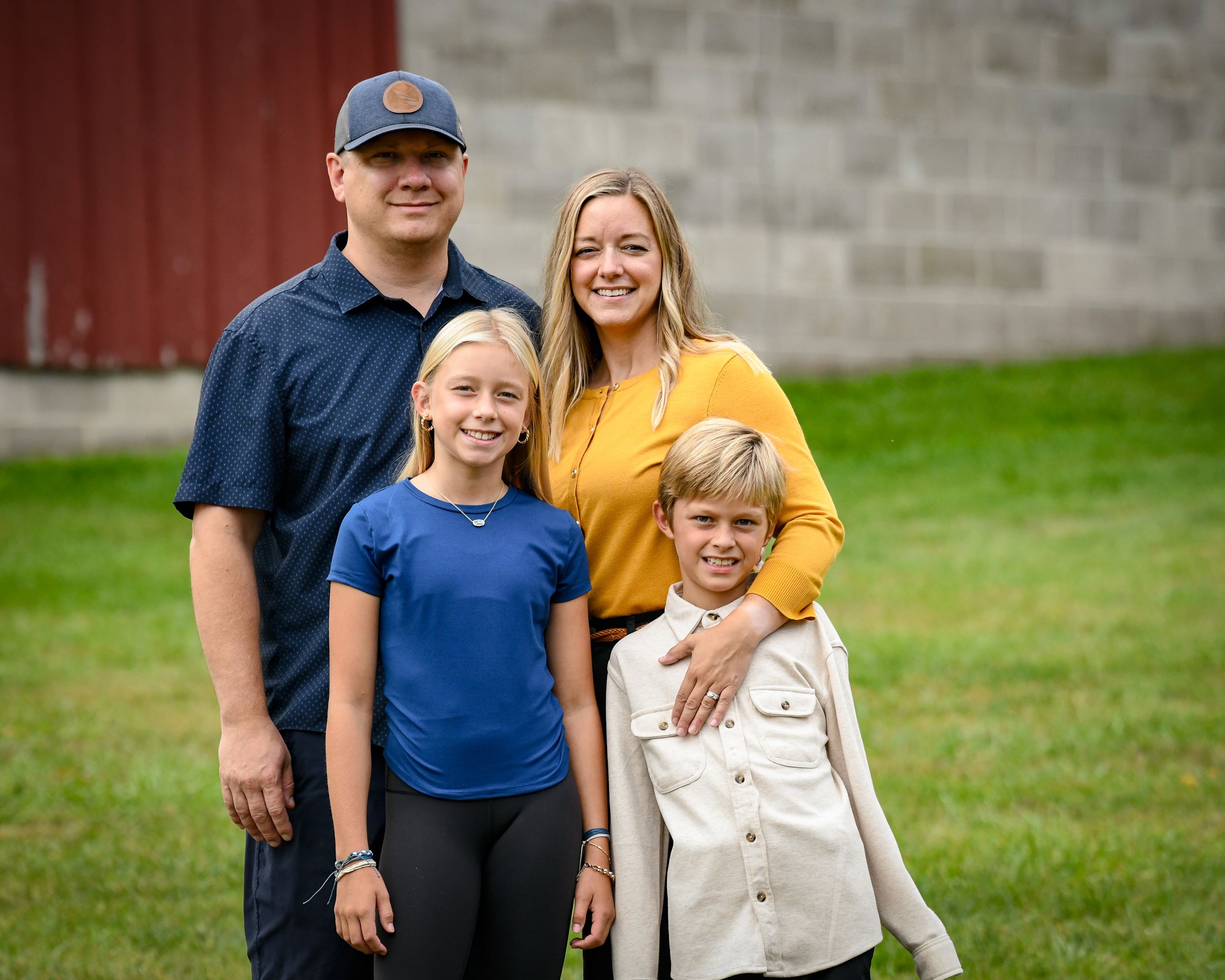 Family of four smiling outdoors near a red barn on a green lawn during a fall photo session.