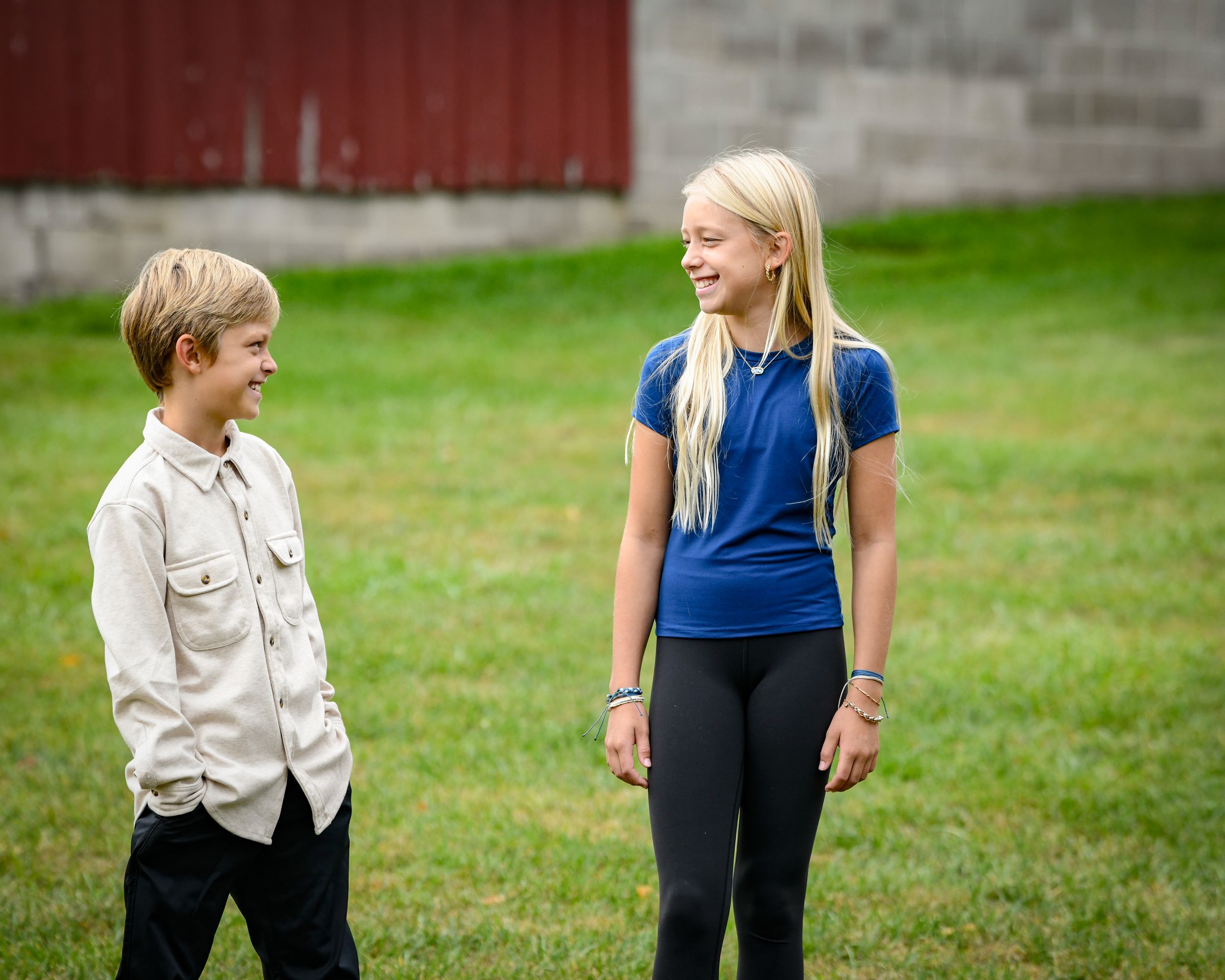 Two children, a boy and a girl with long blonde hair, smiling and talking outdoors on green grass.