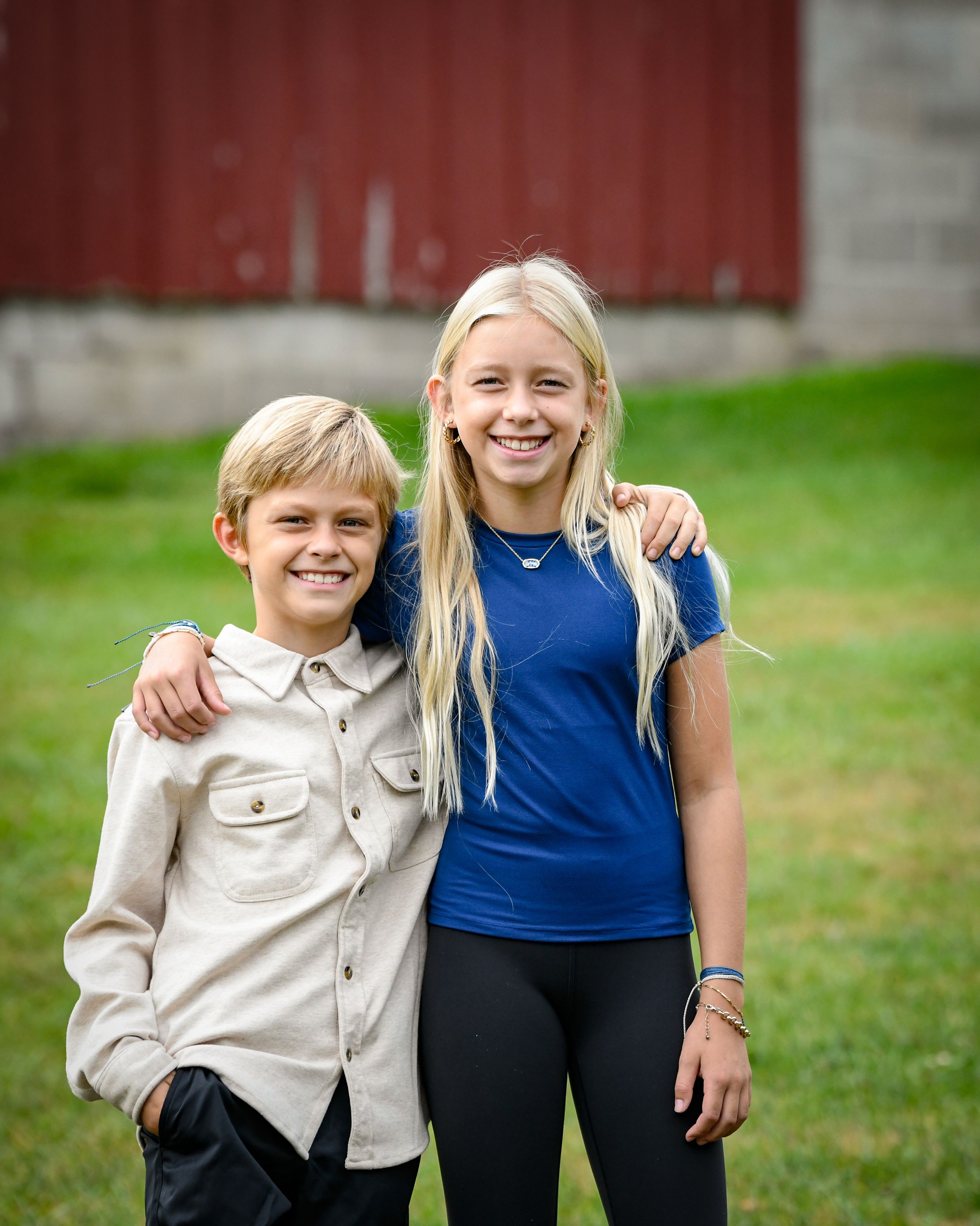 Two smiling blonde children pose together outdoors near a red barn on a green lawn.