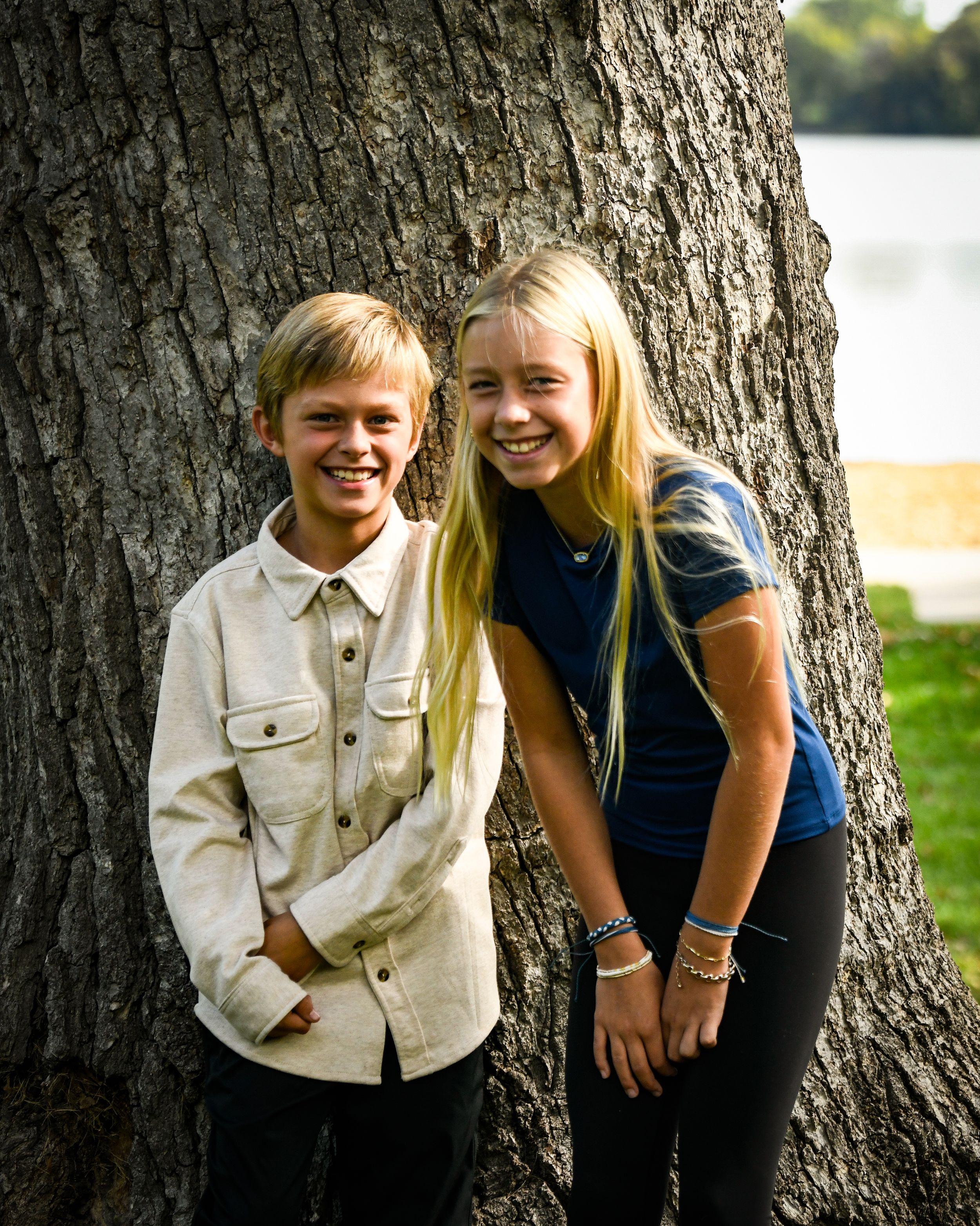 Two smiling kids leaning against a large tree trunk outdoors, a boy in a cream shirt and a girl with long blonde hair.