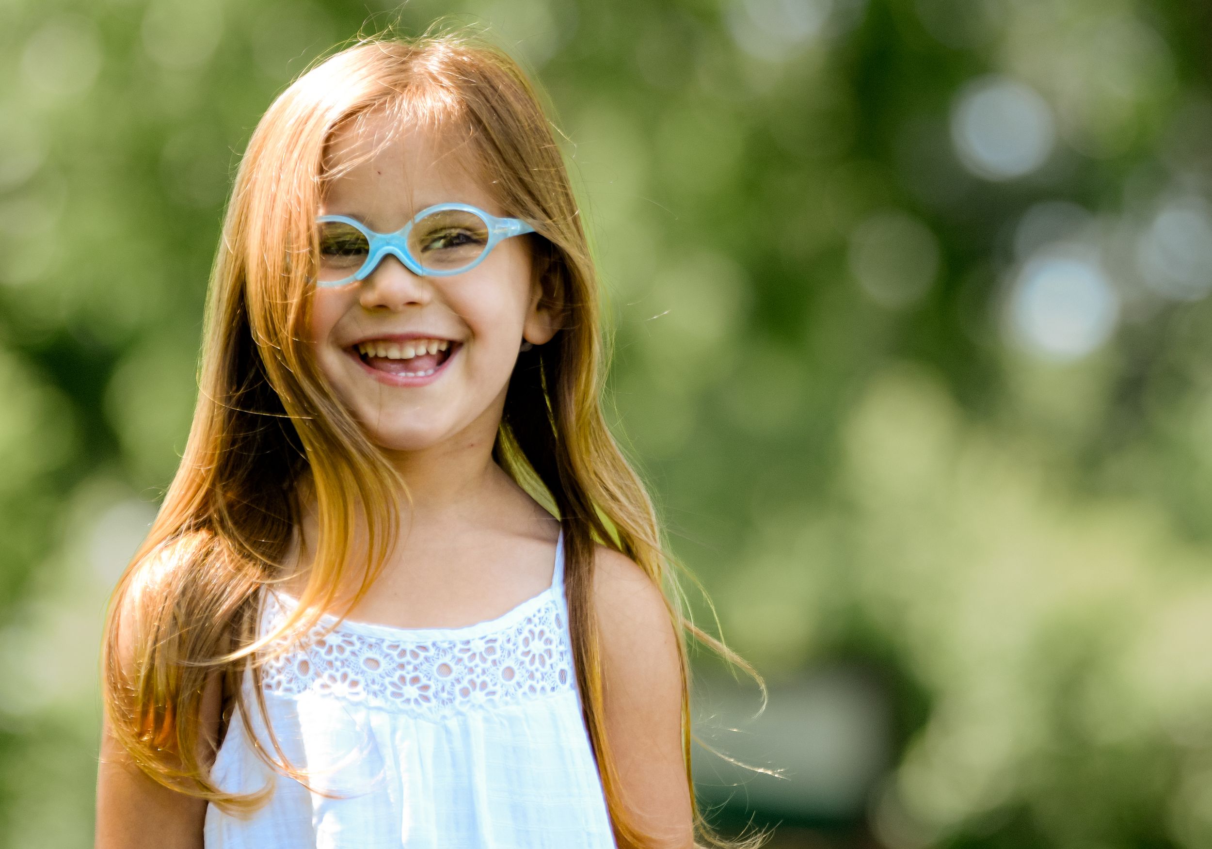 Smiling young girl with long red hair wearing blue glasses outdoors in a sunny green park setting.