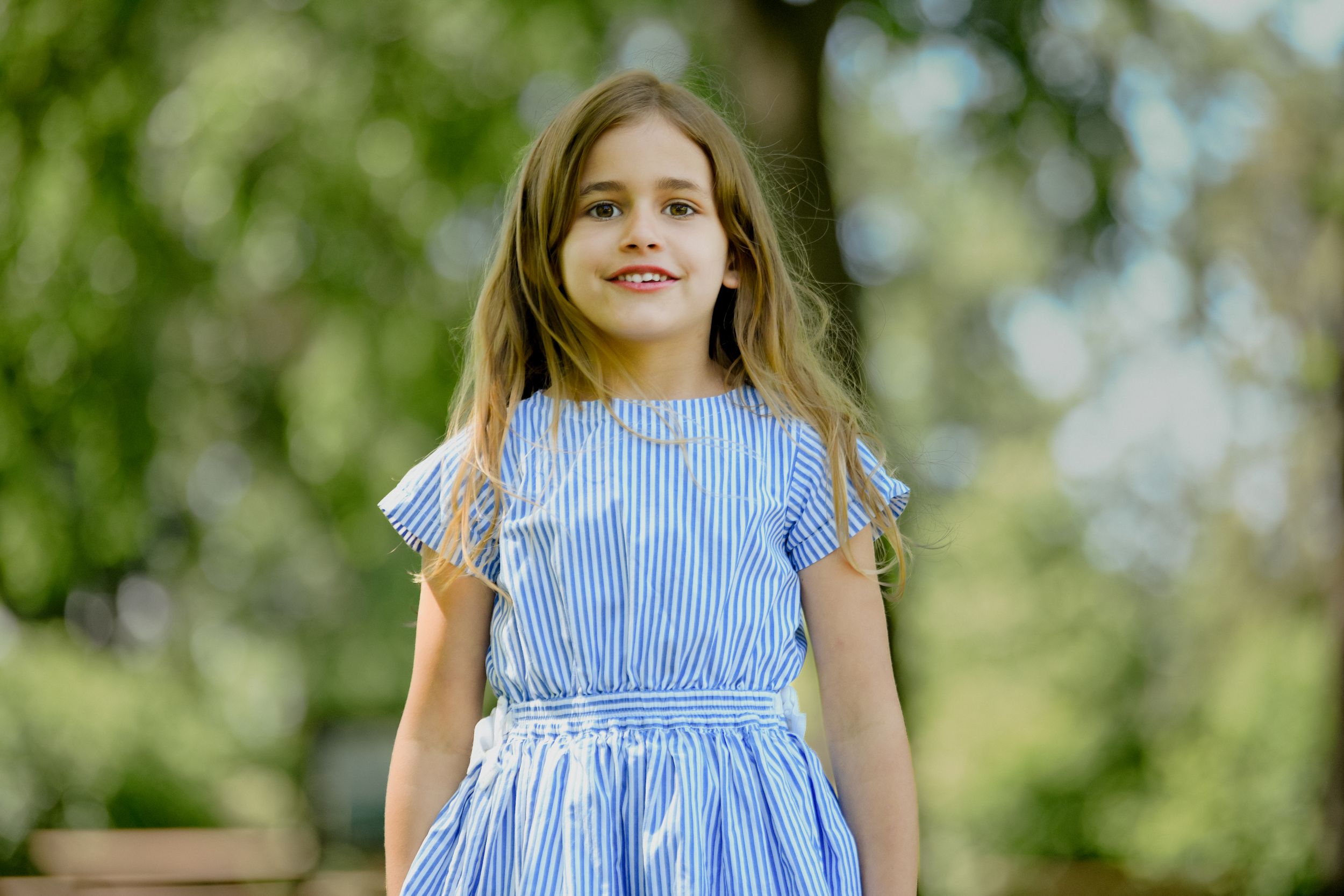 Young girl in blue striped dress smiling outdoors in a green park setting.