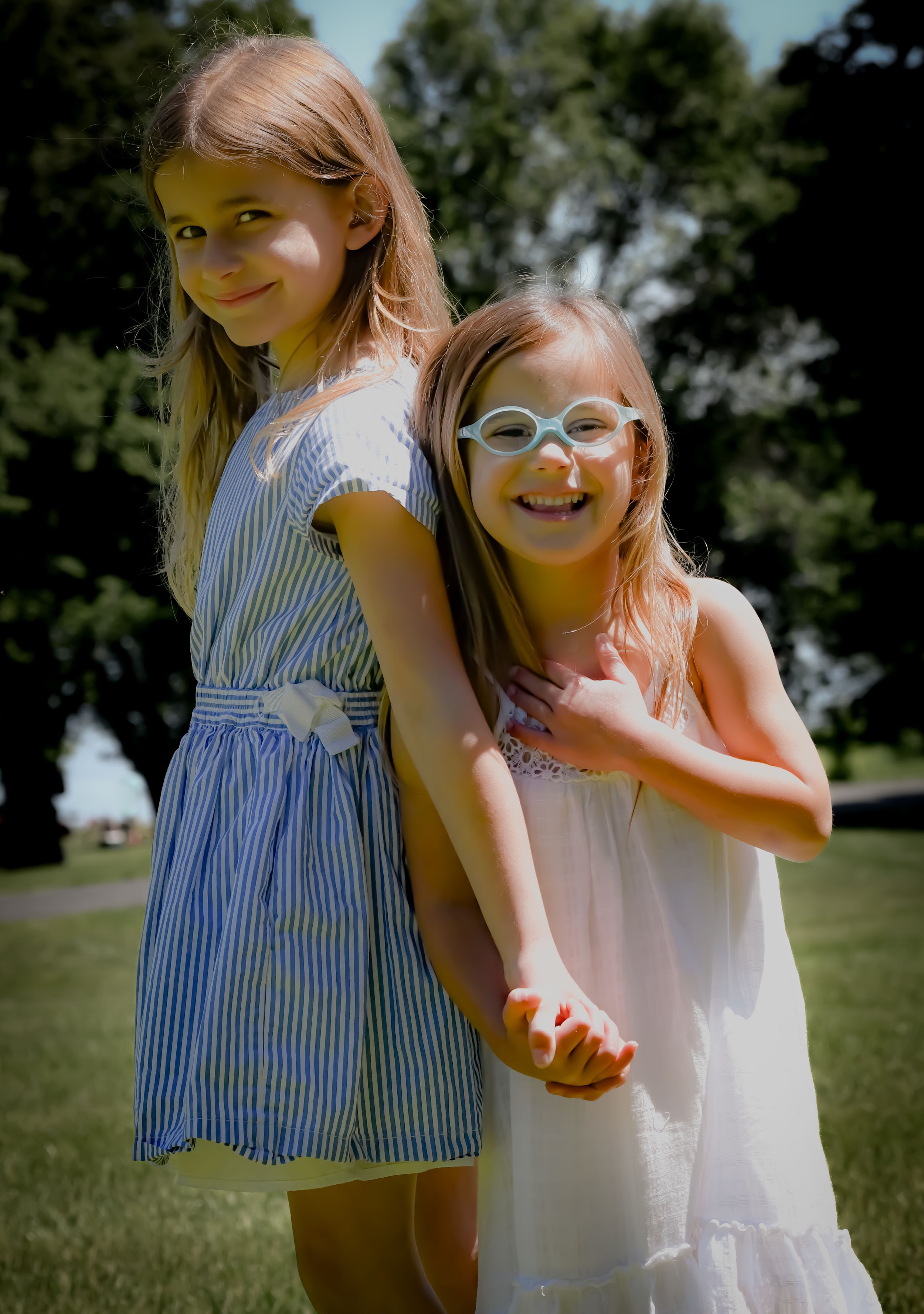 Two young girls smiling outdoors on a sunny day, one in a blue striped dress and one in white wearing glasses.