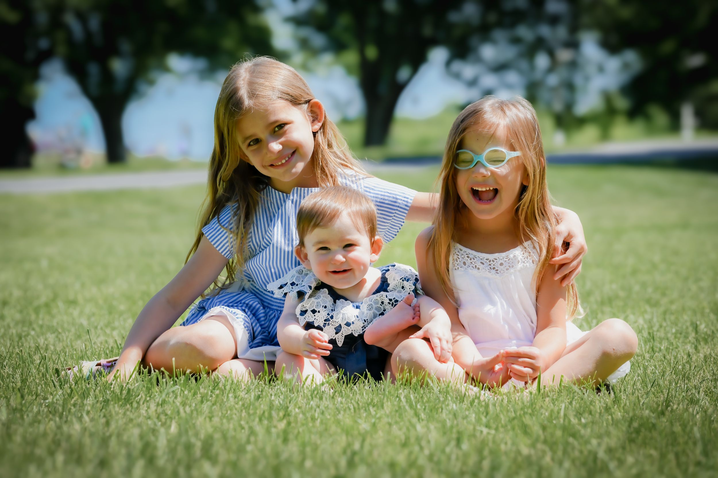 Three happy children sitting on green grass in a sunny park, two older girls smiling with a baby between them.