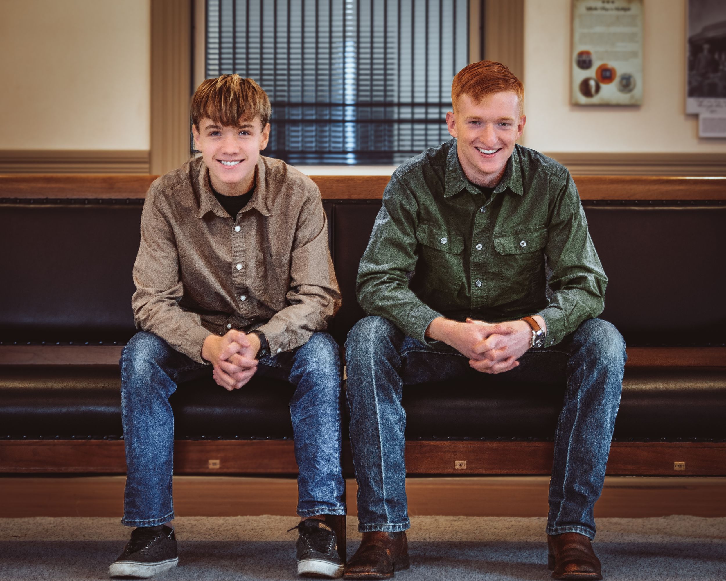 Two young men in button-up shirts and jeans sitting together smiling on a bench indoors.