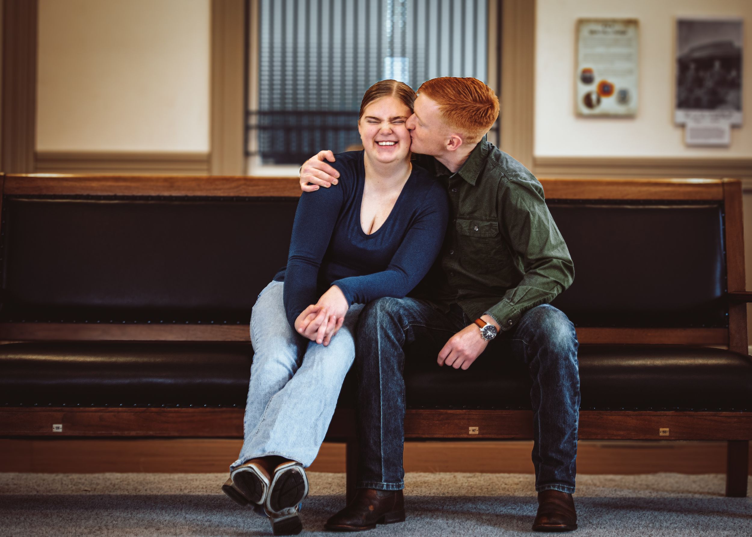 A couple sits on a bench, smiling and sharing a tender moment, with the man kissing the woman's cheek.