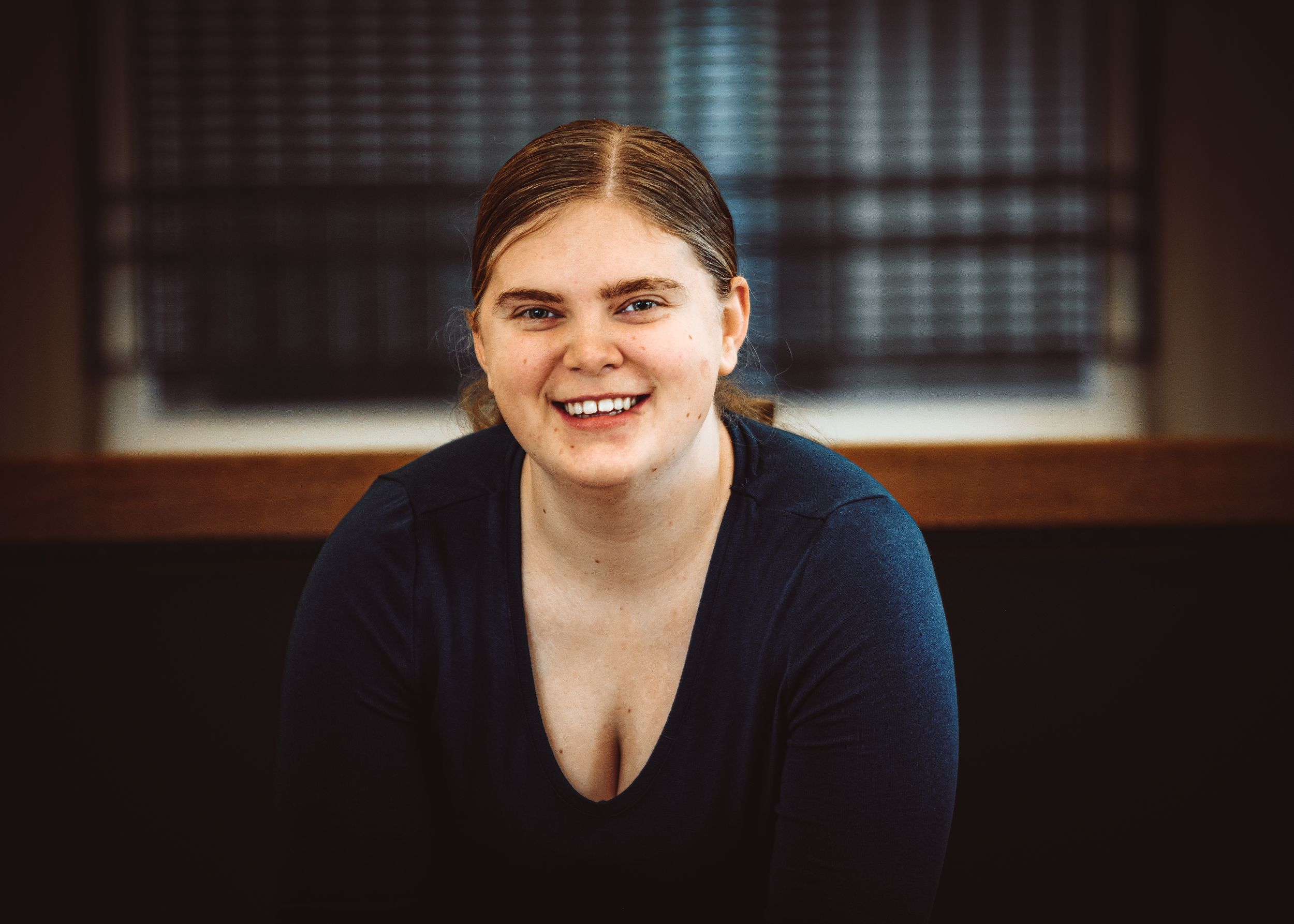 Smiling young woman with brown hair in navy top, seated indoors with warm bokeh background lighting.