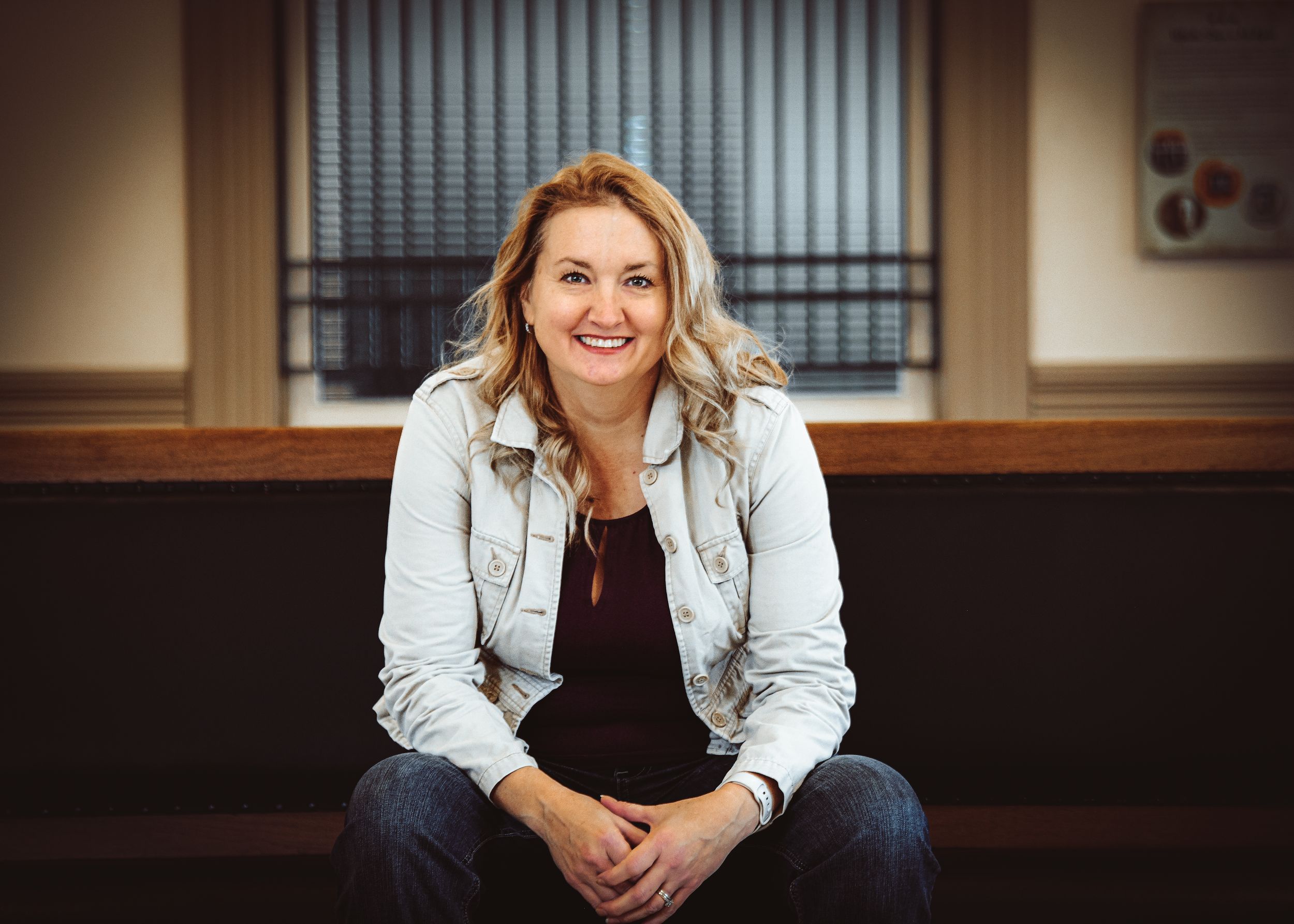 Smiling blonde woman in white denim jacket sitting indoors, posing confidently in a warmly lit modern space.