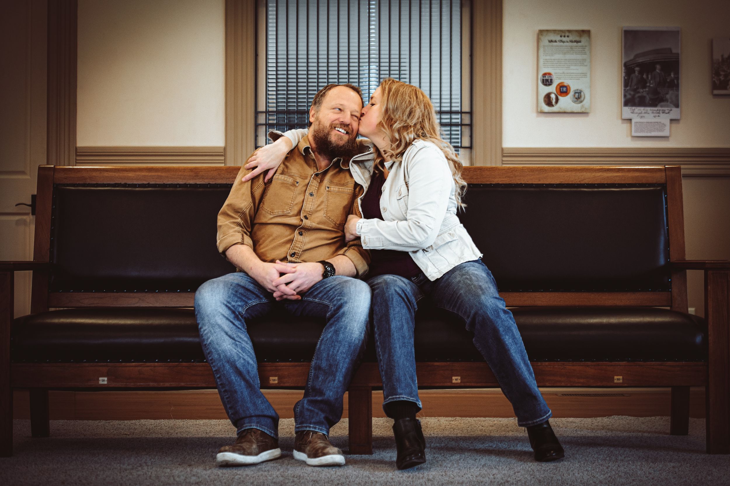 A couple sits on a wooden bench, sharing a tender moment as the woman whispers to the smiling bearded man.