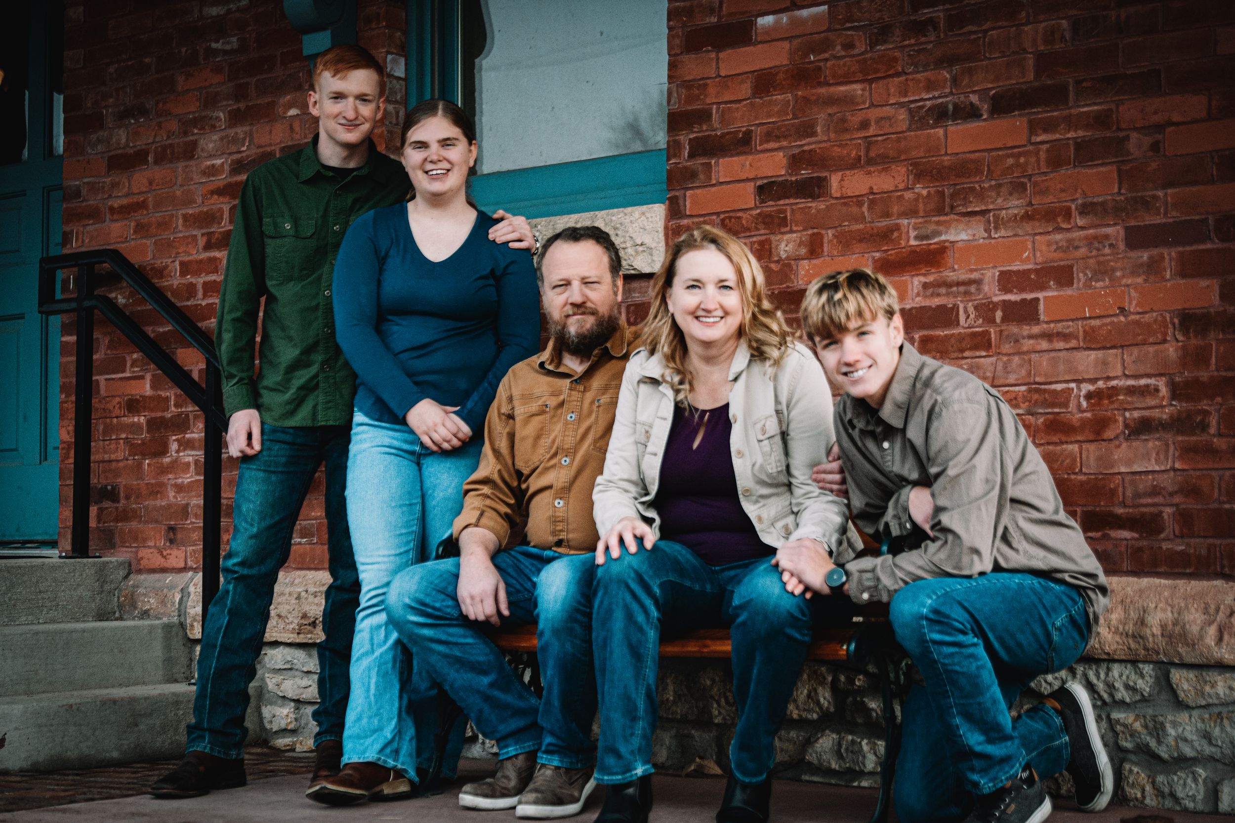 Family of five smiling together on brick building steps, casually dressed in jeans and colorful tops.