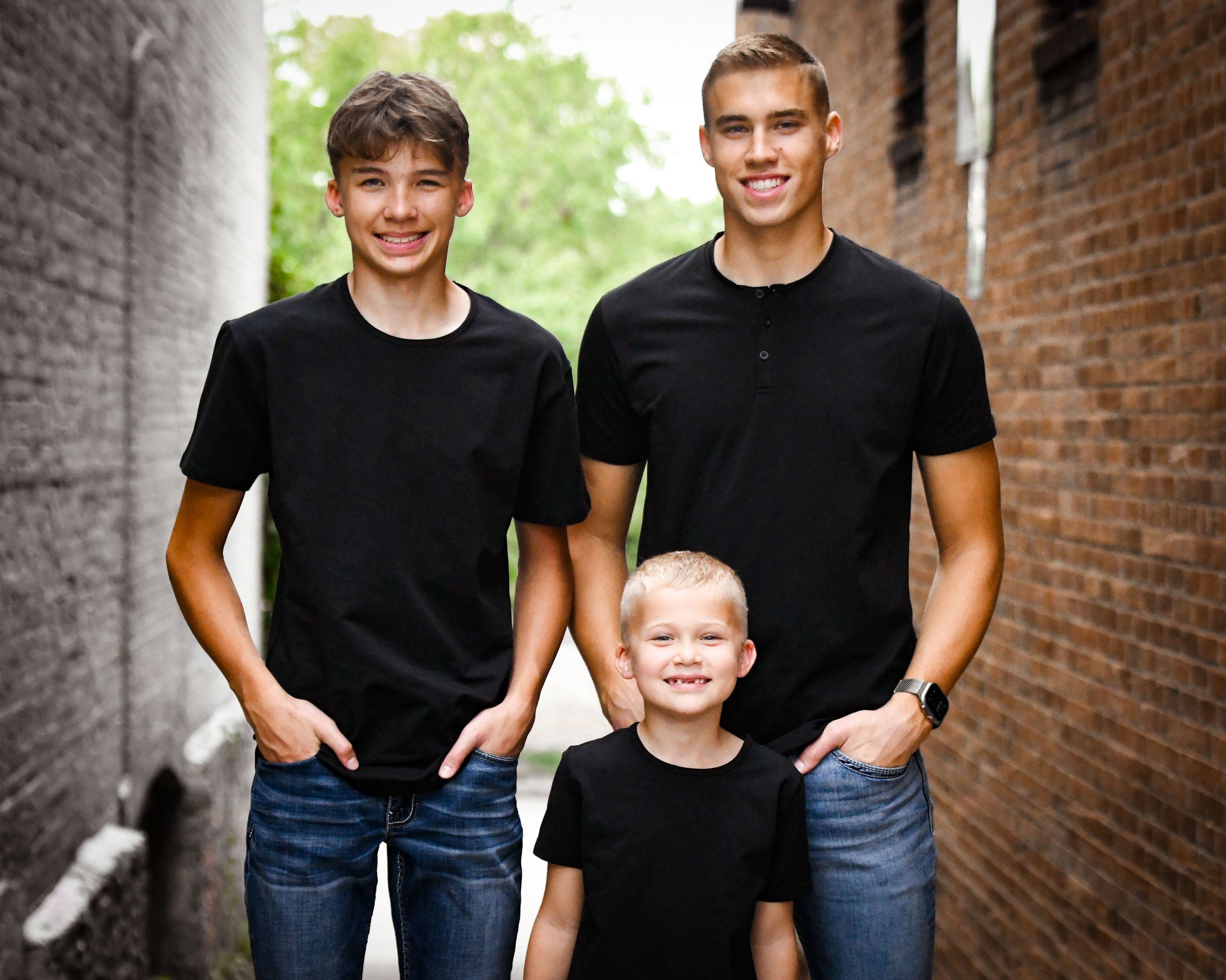 Three boys in matching black t-shirts and jeans pose in a brick alley, two teenagers flanking a young blonde child.