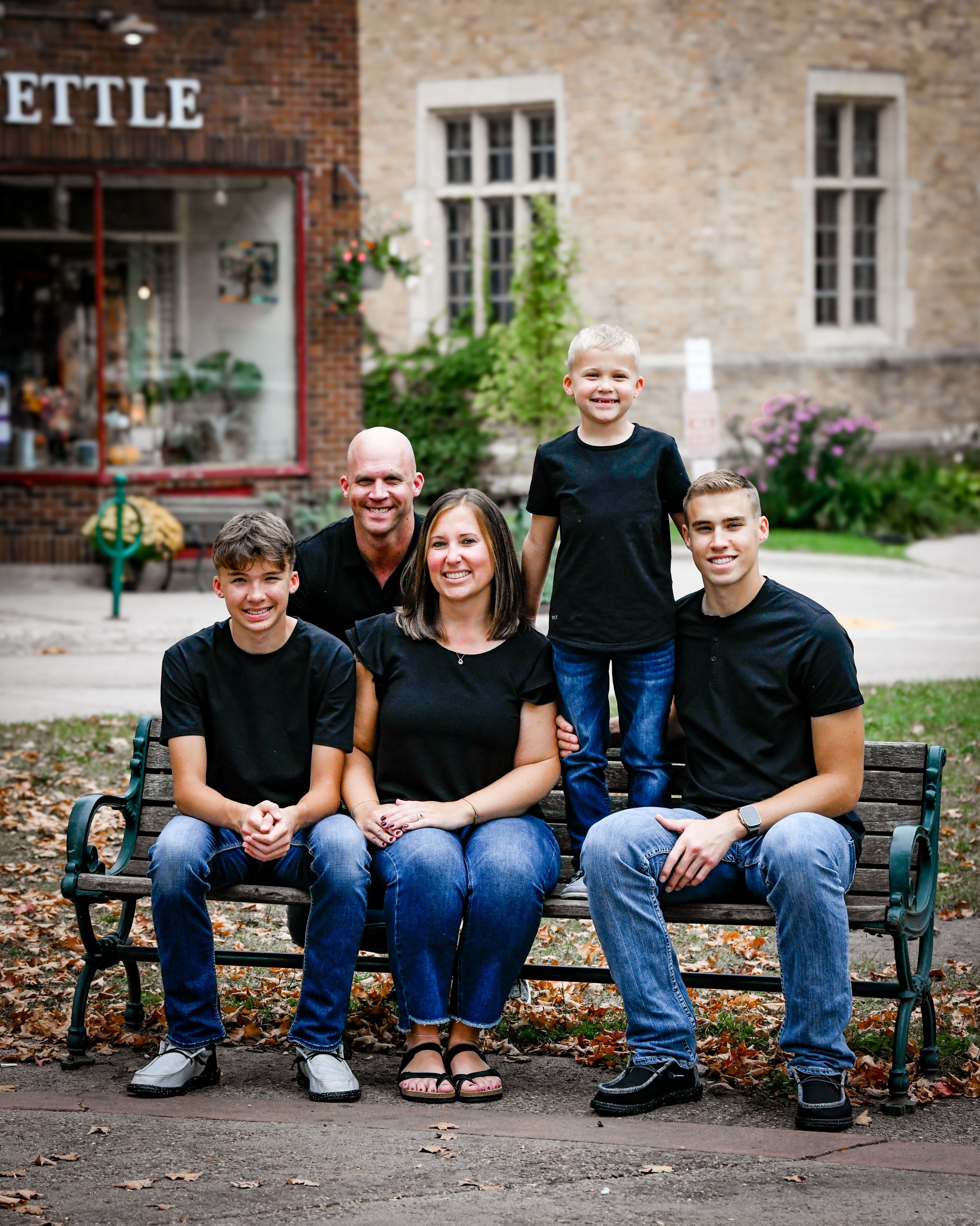 Family of five posing on a bench outdoors, wearing matching black tops and jeans, smiling in front of a brick building.