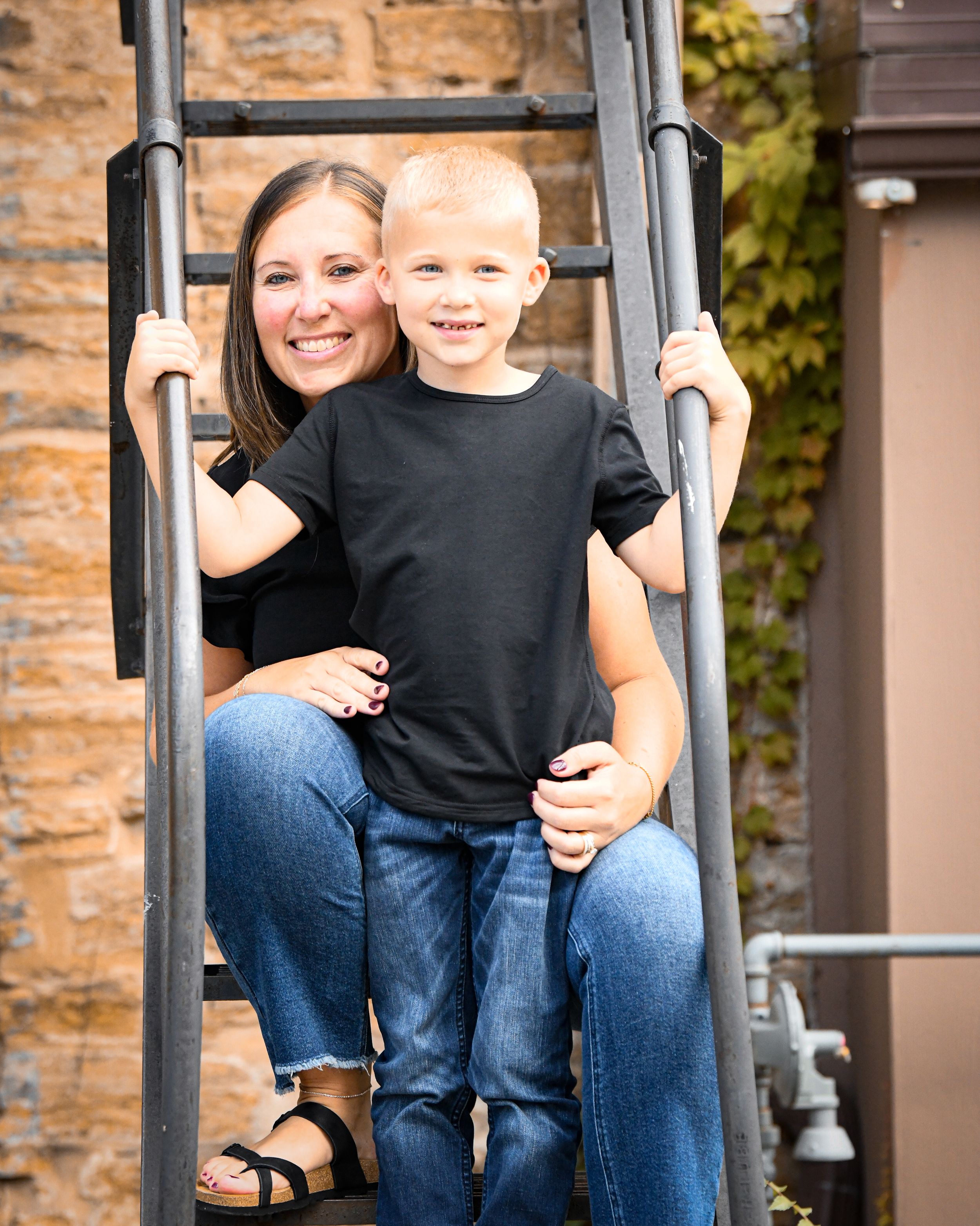 A smiling woman and young boy in matching black shirts and jeans pose together on a metal ladder outdoors.