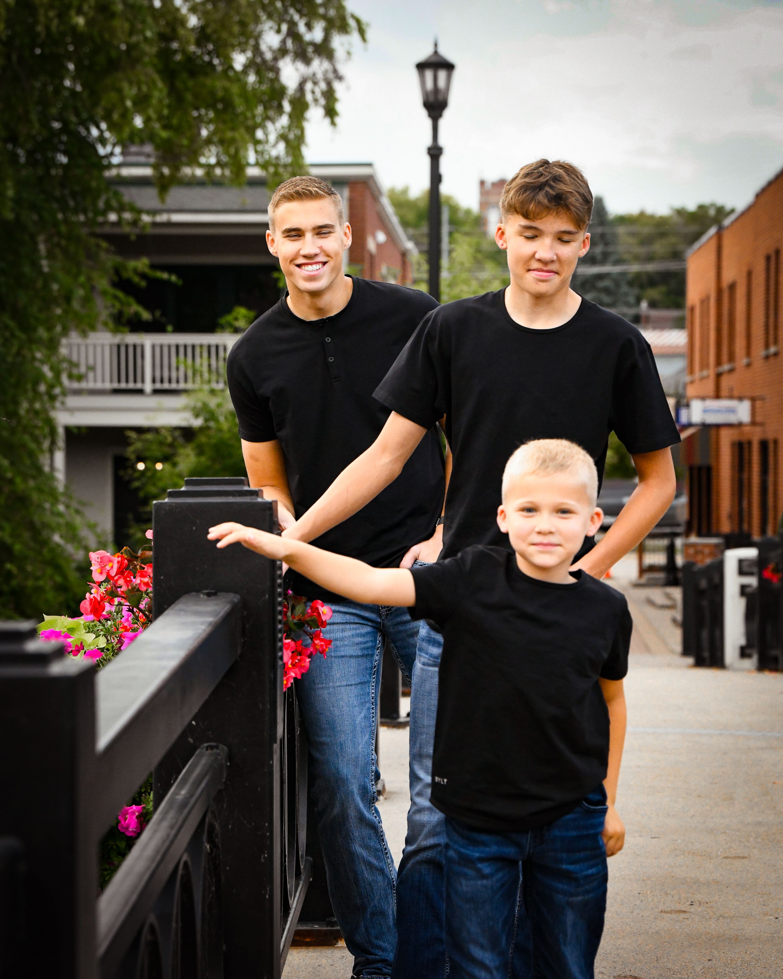 Three boys in matching black shirts pose on a charming town bridge, with the oldest two standing behind the youngest.