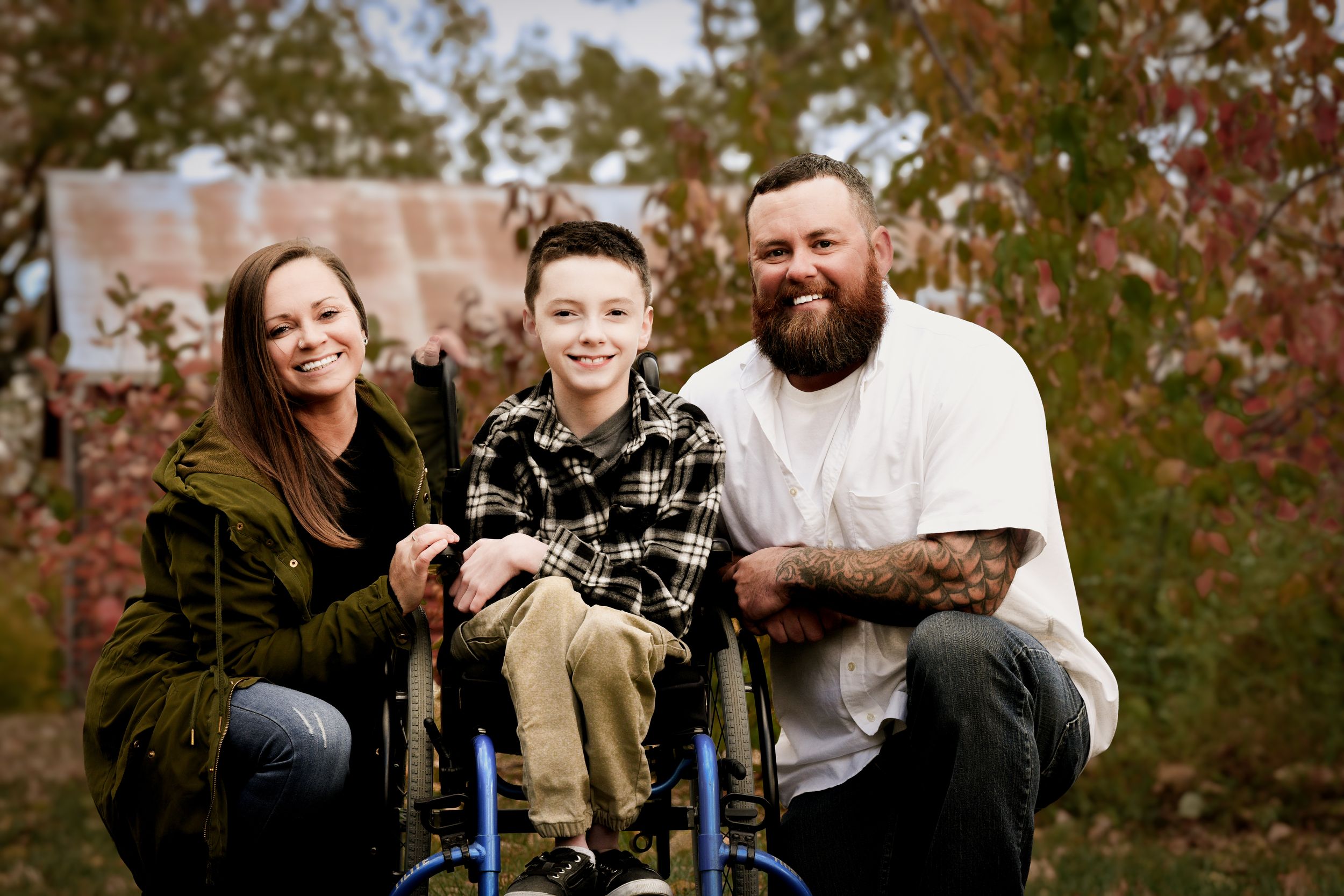 Smiling family with boy in blue wheelchair posing outdoors among colorful autumn foliage.
