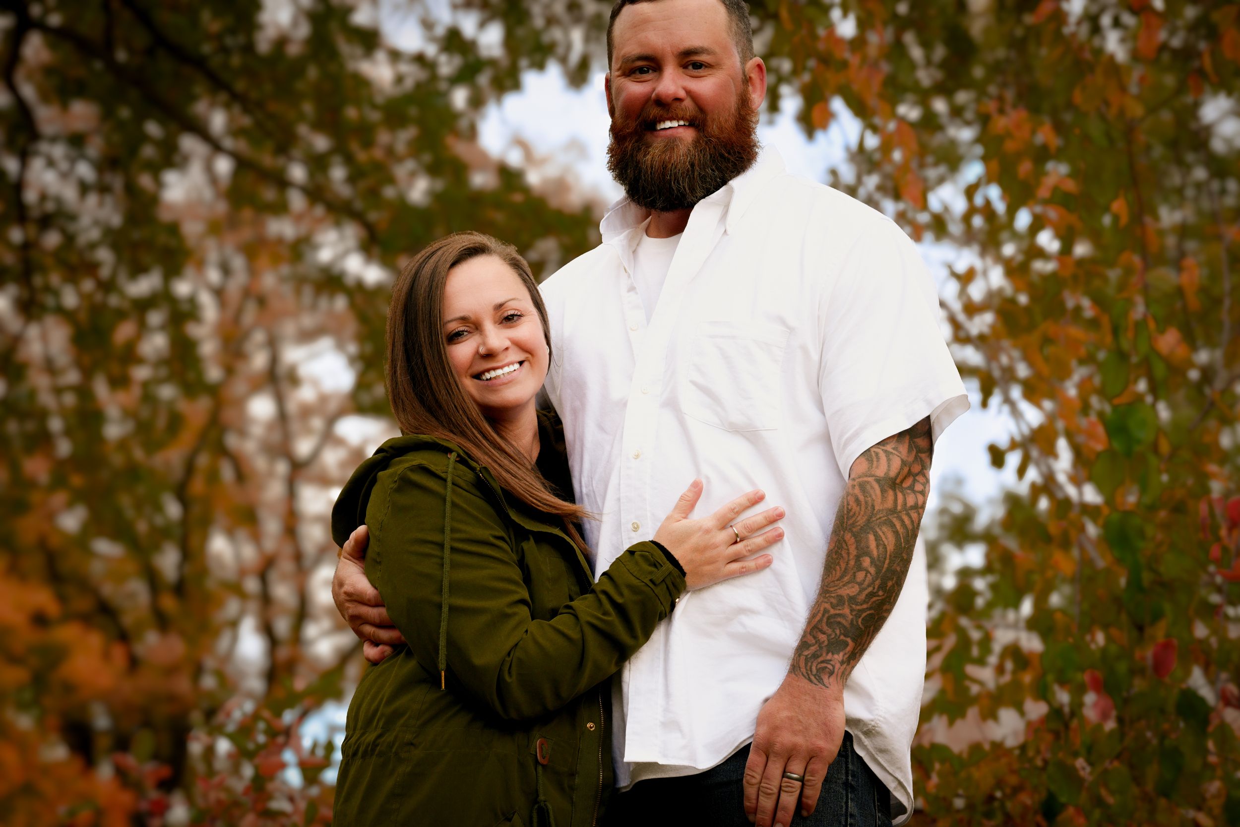 Smiling couple embracing outdoors surrounded by vibrant fall foliage, man with tattooed arm.