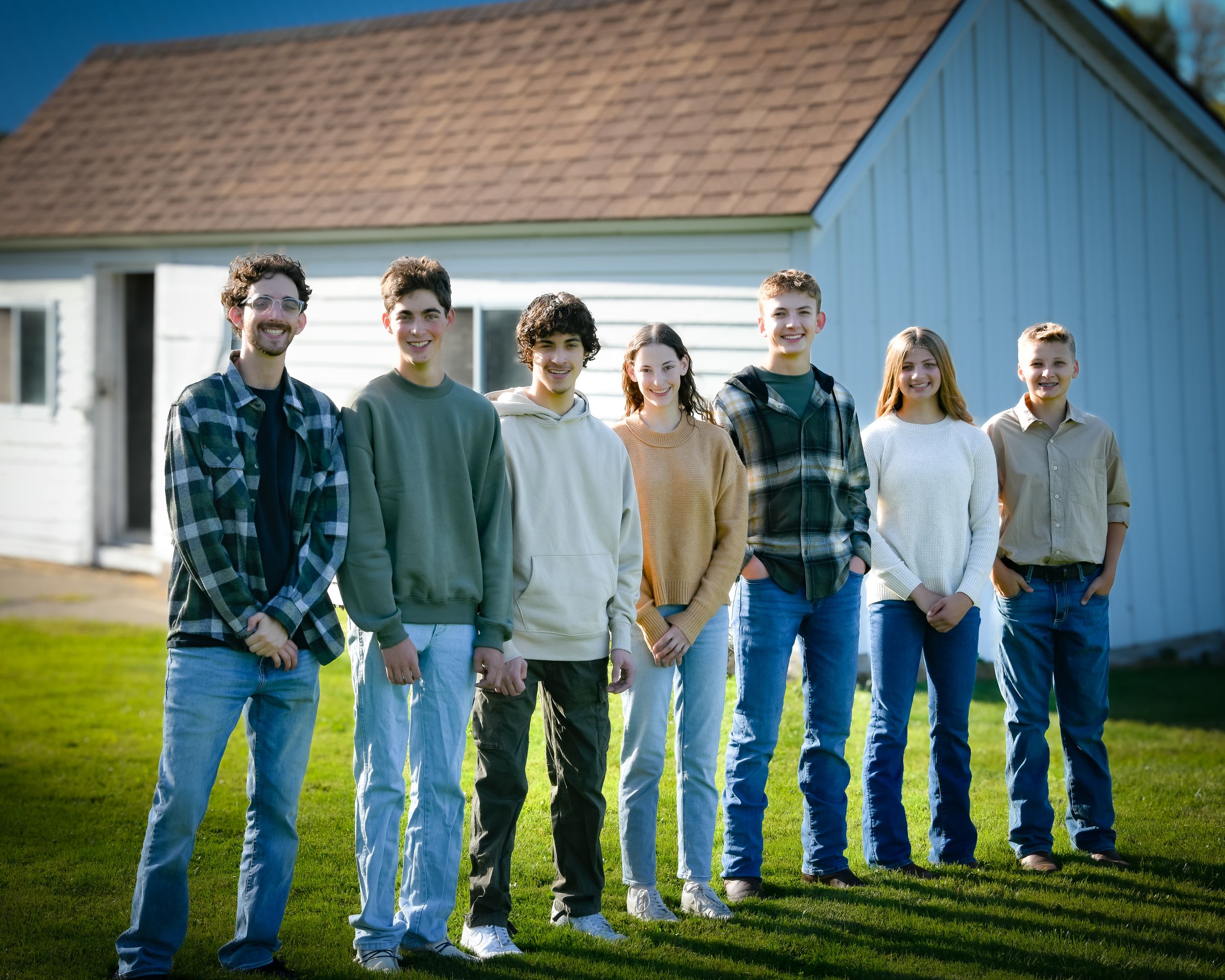 Seven young adults smiling and posing together outdoors on green grass in front of a white farmhouse building.