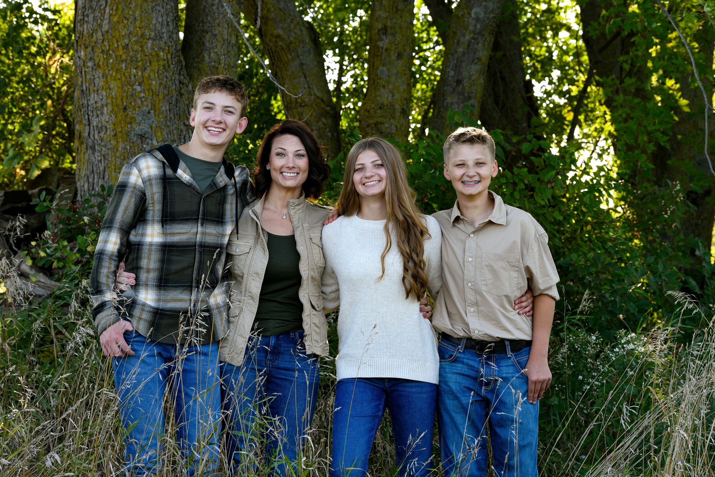 Family of four smiling together outdoors in front of large trees in a natural wooded setting.