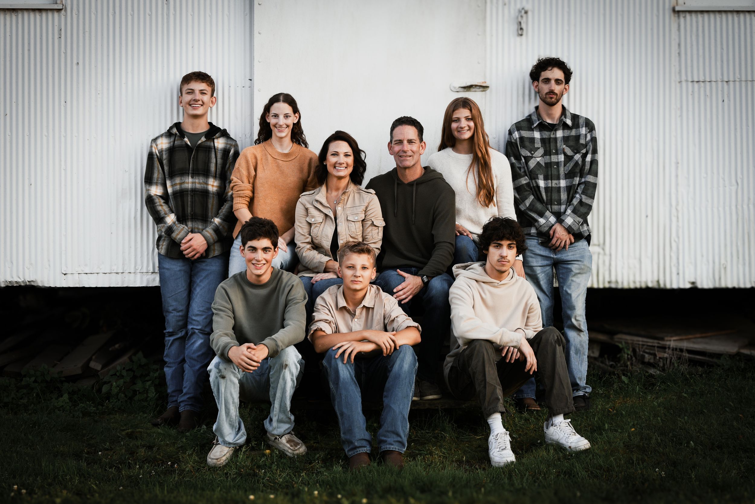 Large family of nine posing together outdoors in front of a white barn, smiling for a group portrait.