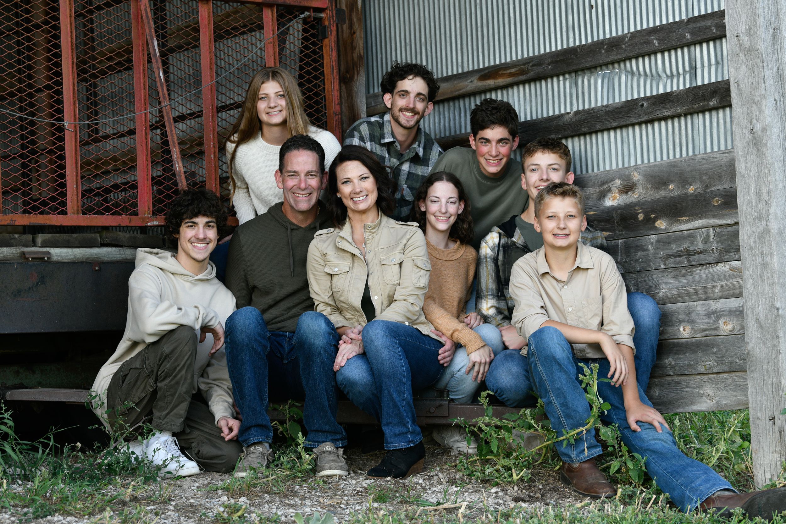 Large family group smiling together outdoors near a rustic barn, including adults and children with a small dog.