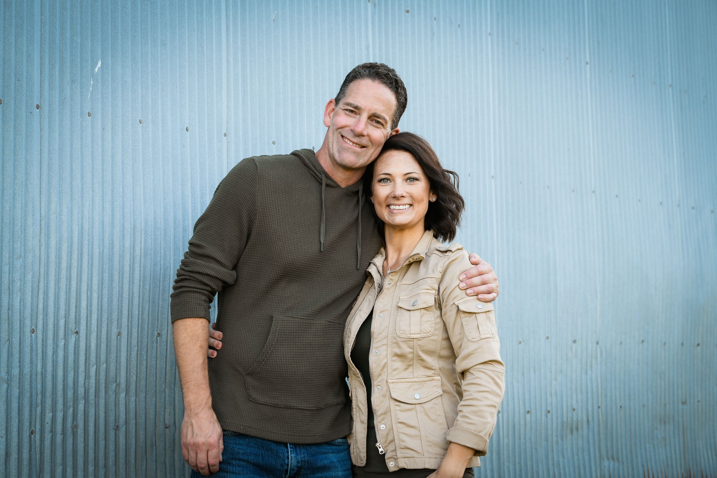 A smiling man and woman pose together against a corrugated metal wall in casual outdoor clothing.