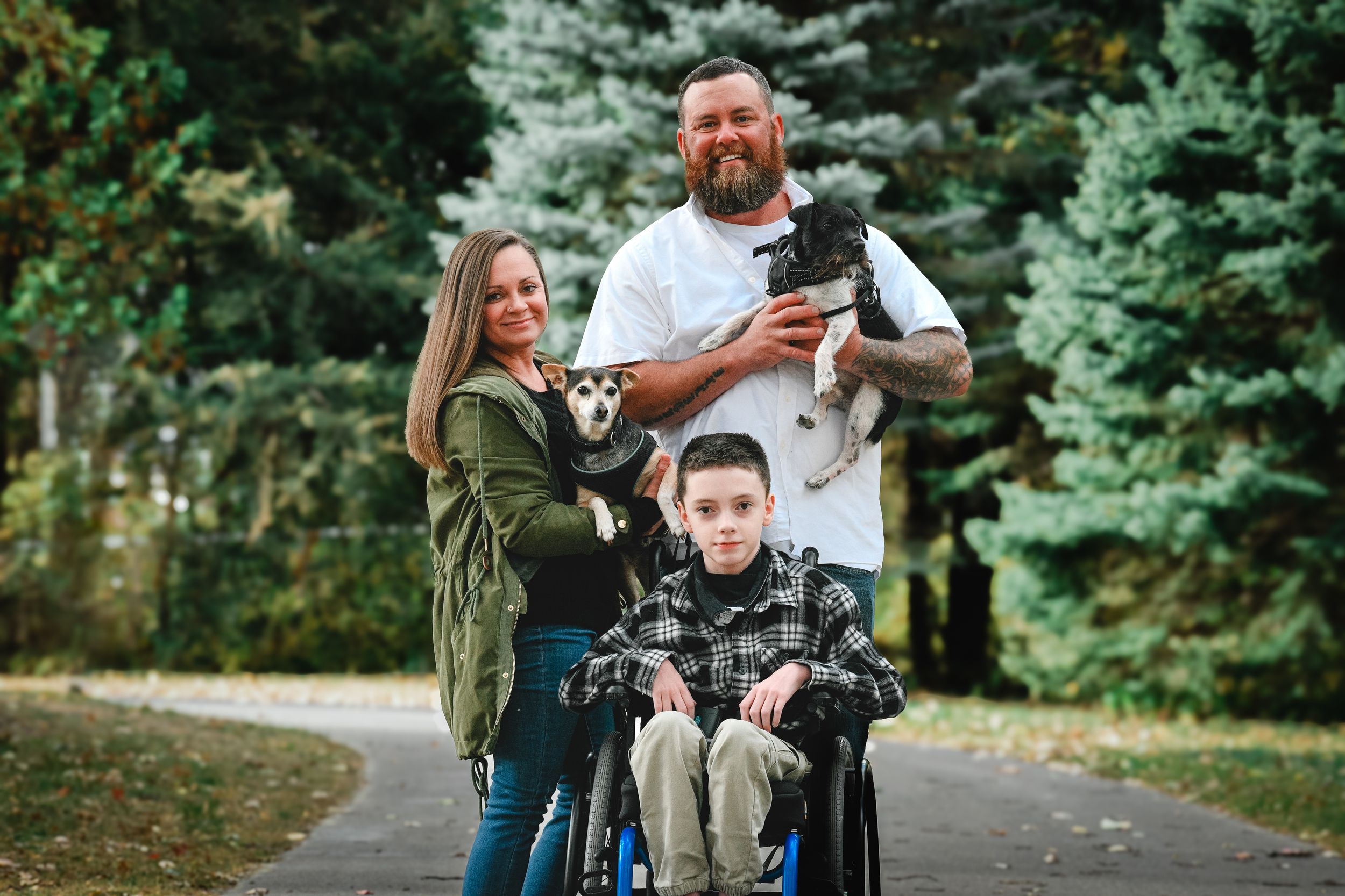 Family with boy in wheelchair, woman, bearded man holding dogs, posing on park path surrounded by trees.