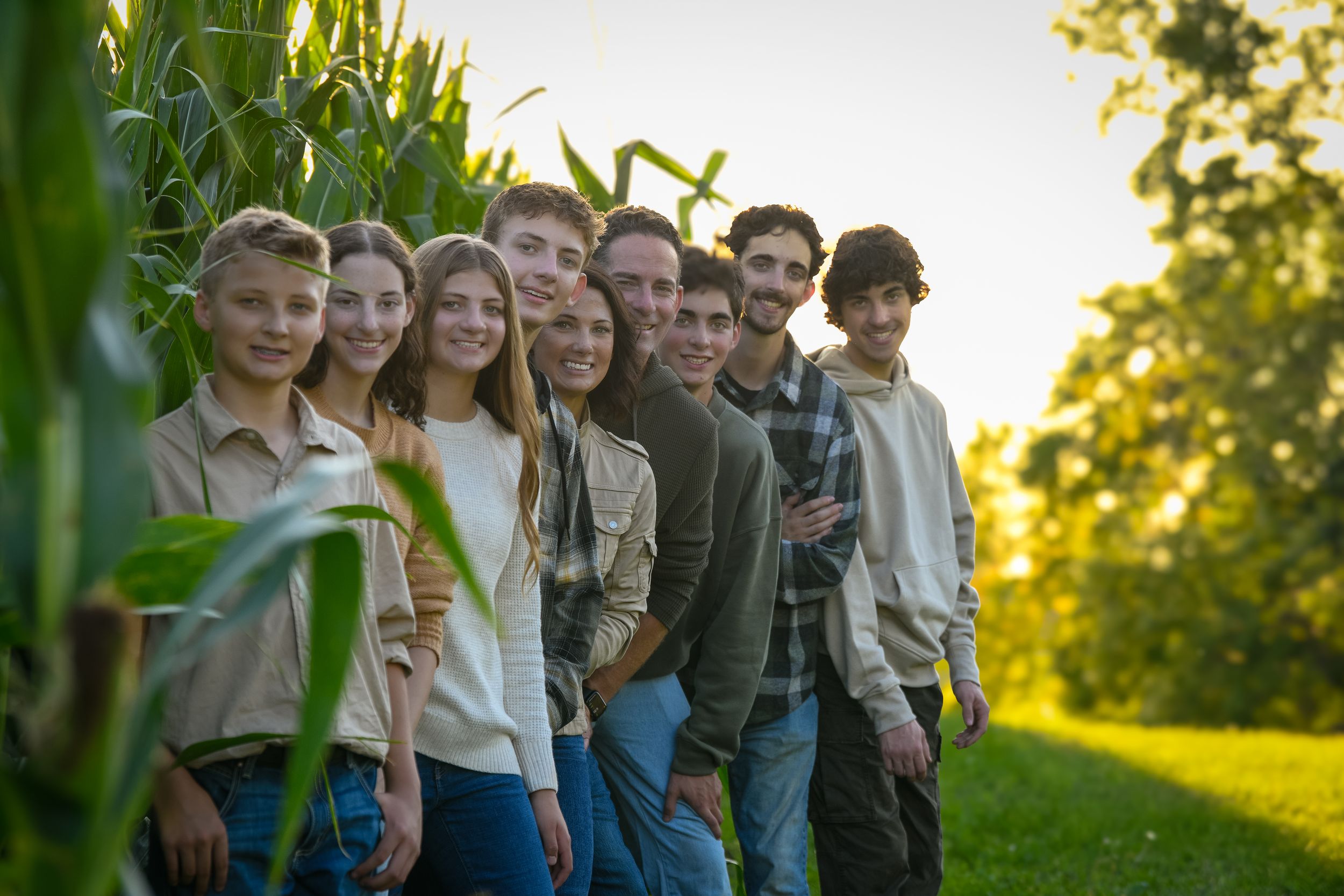 Group of teenagers posing together in a row beside tall green corn stalks during golden hour outdoors.