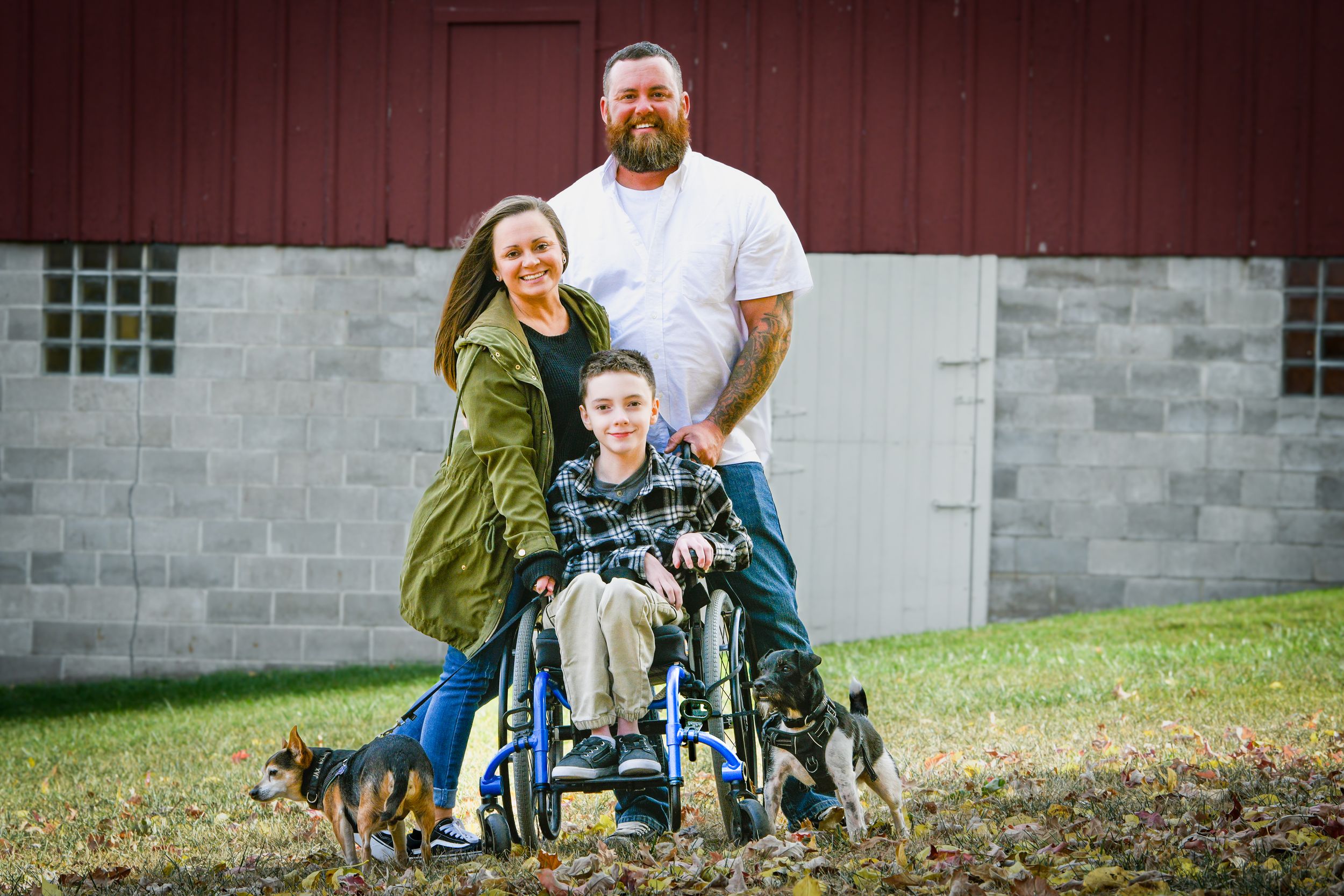 Family portrait with woman, bearded man, boy in wheelchair, and two dogs in front of a red barn.