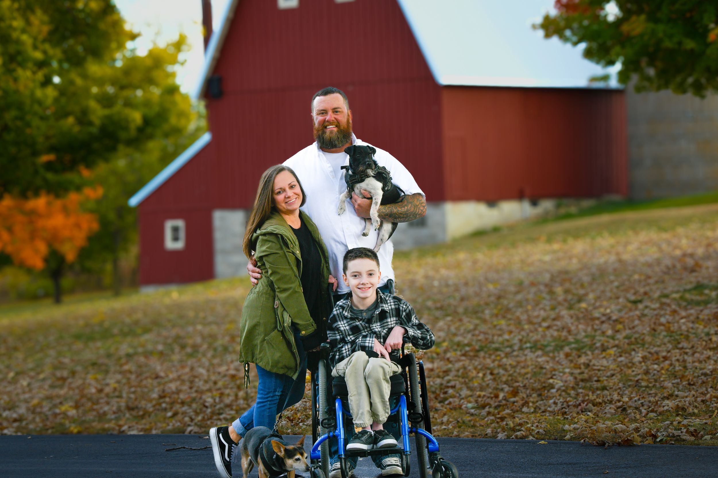 Family portrait with mom, dad, and son in wheelchair smiling outdoors near red barn with fall foliage.