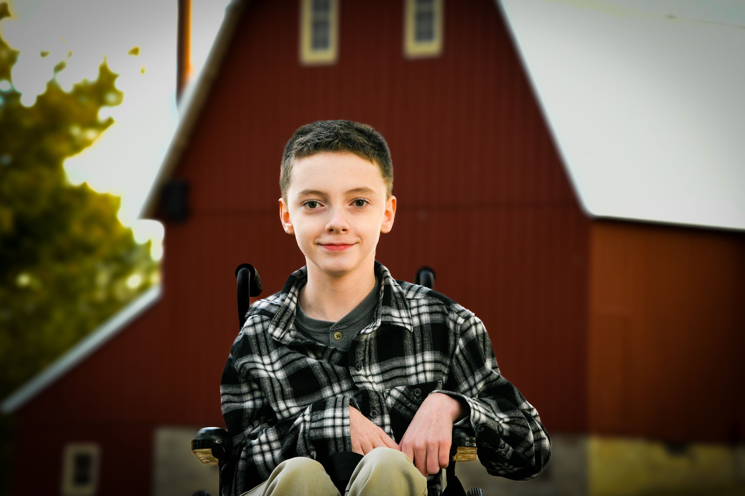 A smiling boy in a wheelchair wearing a plaid shirt poses outdoors in front of a red barn.