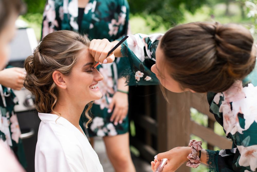 Bridesmaid applies eye makeup to smiling bride wearing white robe, both outdoors with others in floral robes nearby.