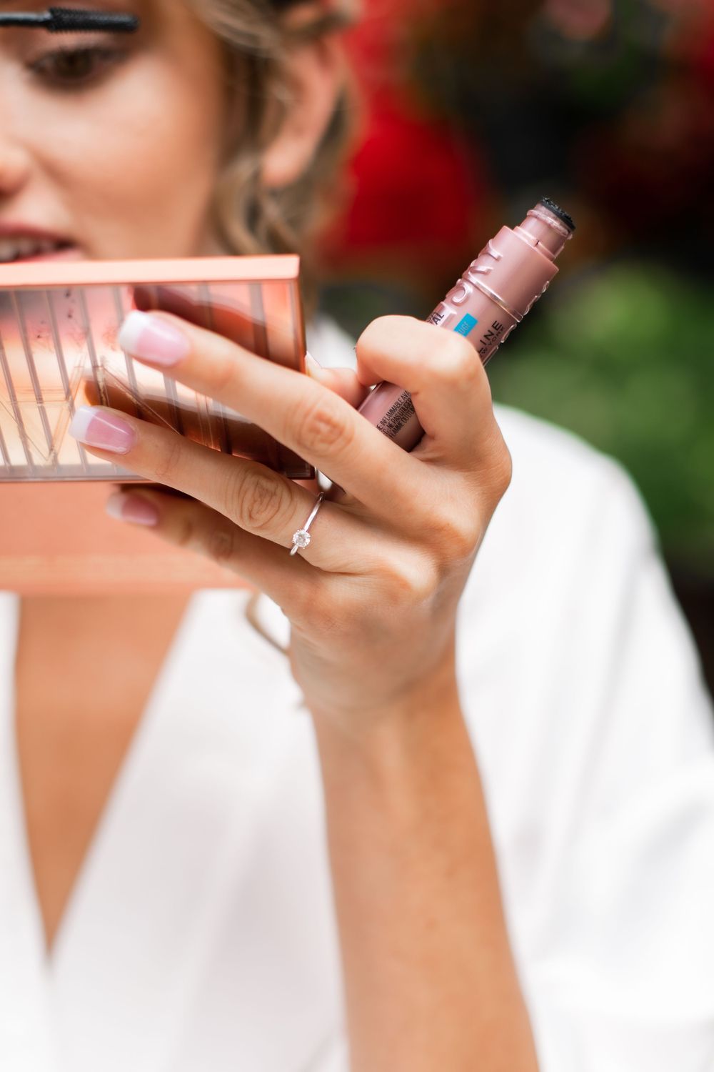 Close-up of bride in white robe holding pink makeup palette and lipstick with delicate engagement ring visible.