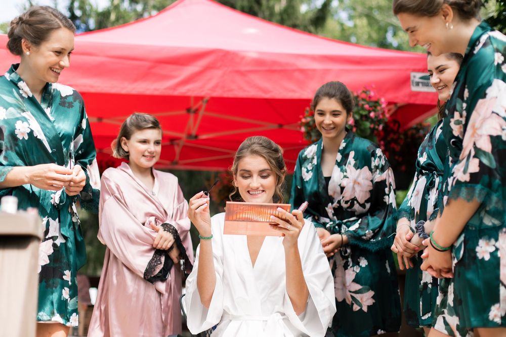 Bride in white robe opens gift surrounded by smiling bridesmaids in green floral robes outdoors near red tent.